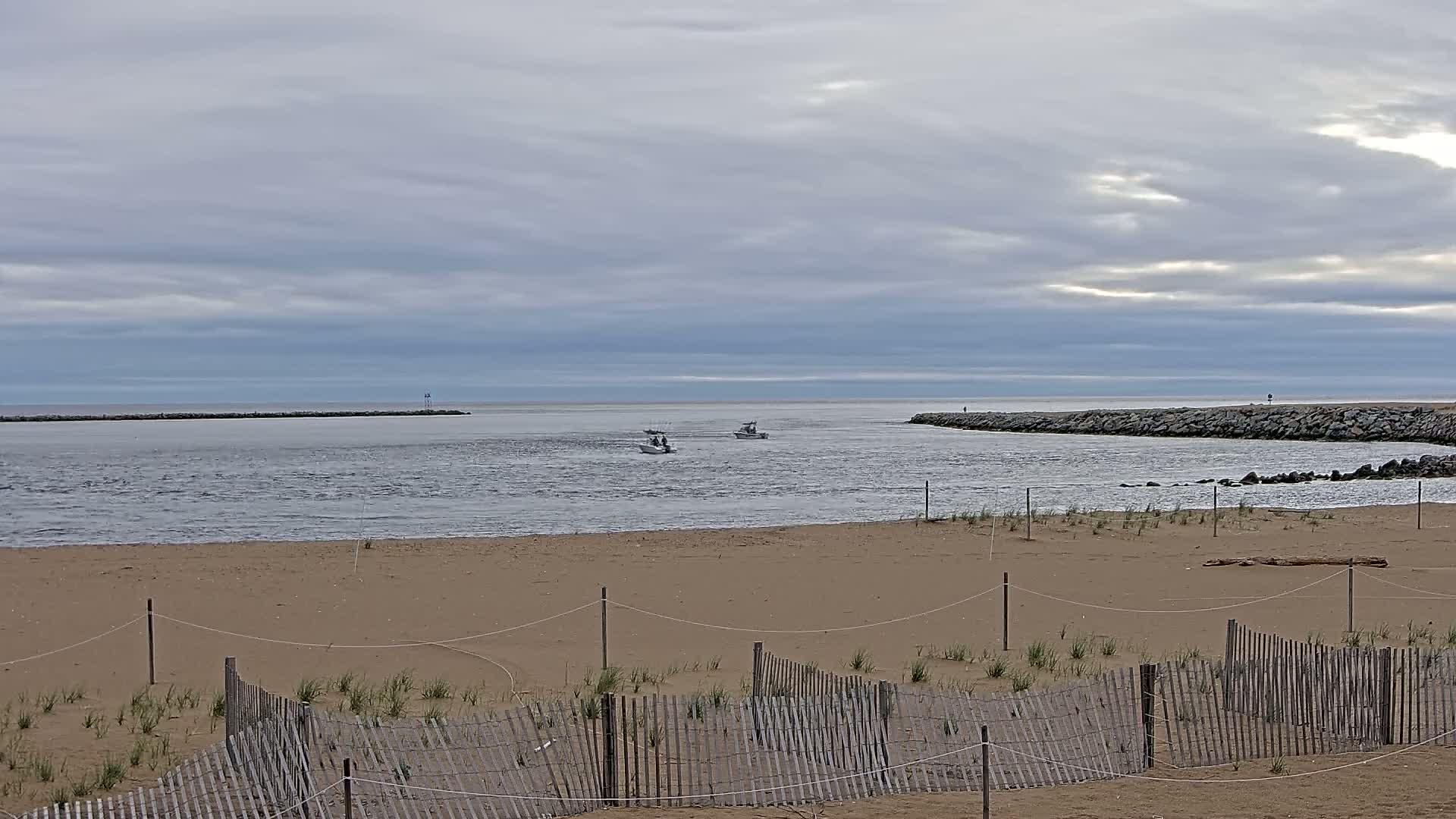 A sandy beach with a wooden fence is shown, overlooking a calm inlet where several small boats are visible under a cloudy sky.