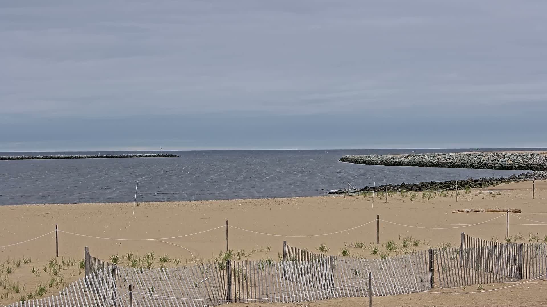 A sandy beach with a low wooden fence is bordered by calm, slightly wavy water and rock jetties under a cloudy sky.