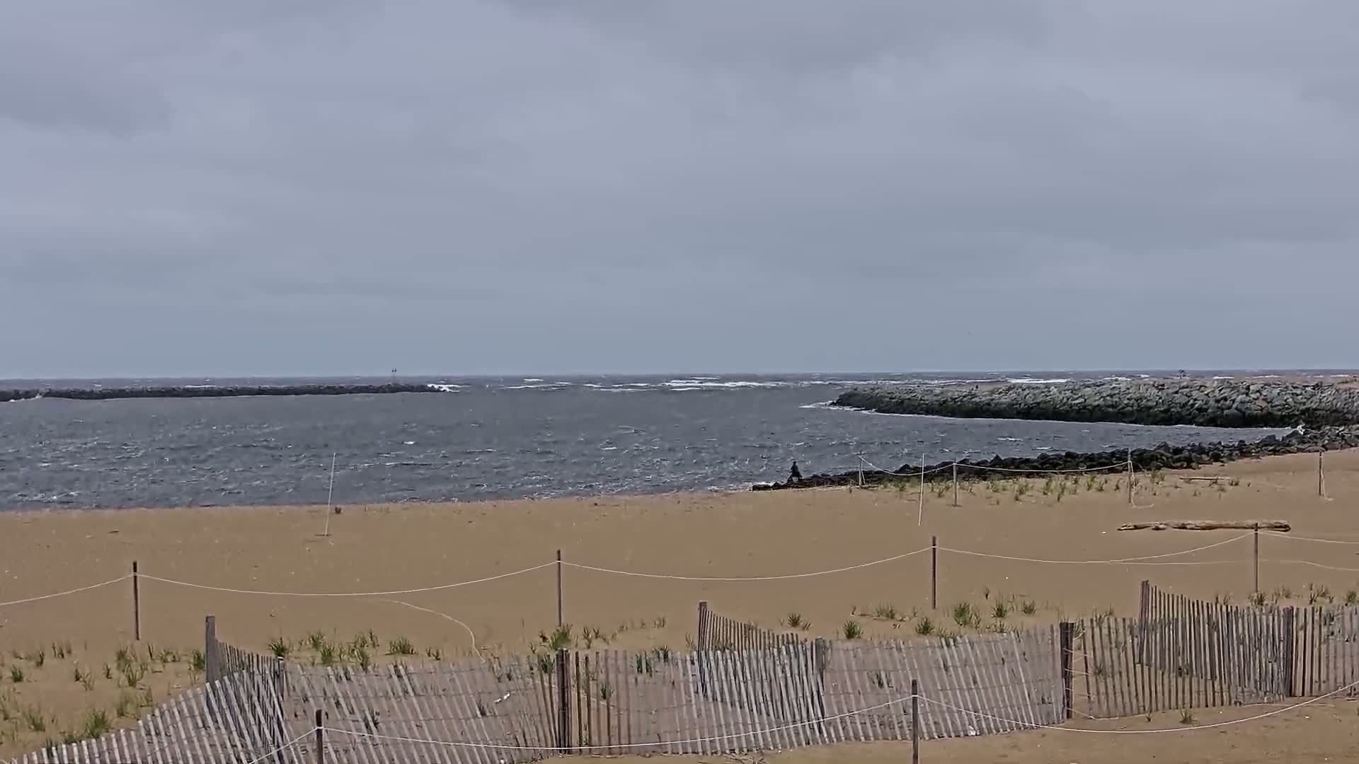 A sandy beach with a wooden fence is shown, overlooking a choppy sea with rocky jetties on a cloudy day.