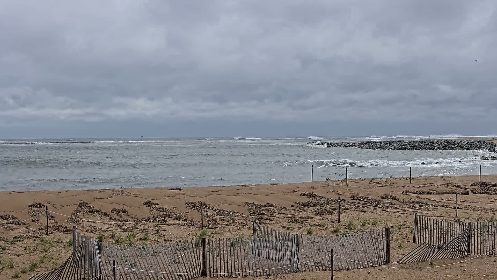 A sandy beach with small wooden fences is shown under a cloudy sky, with a choppy ocean visible in the background.