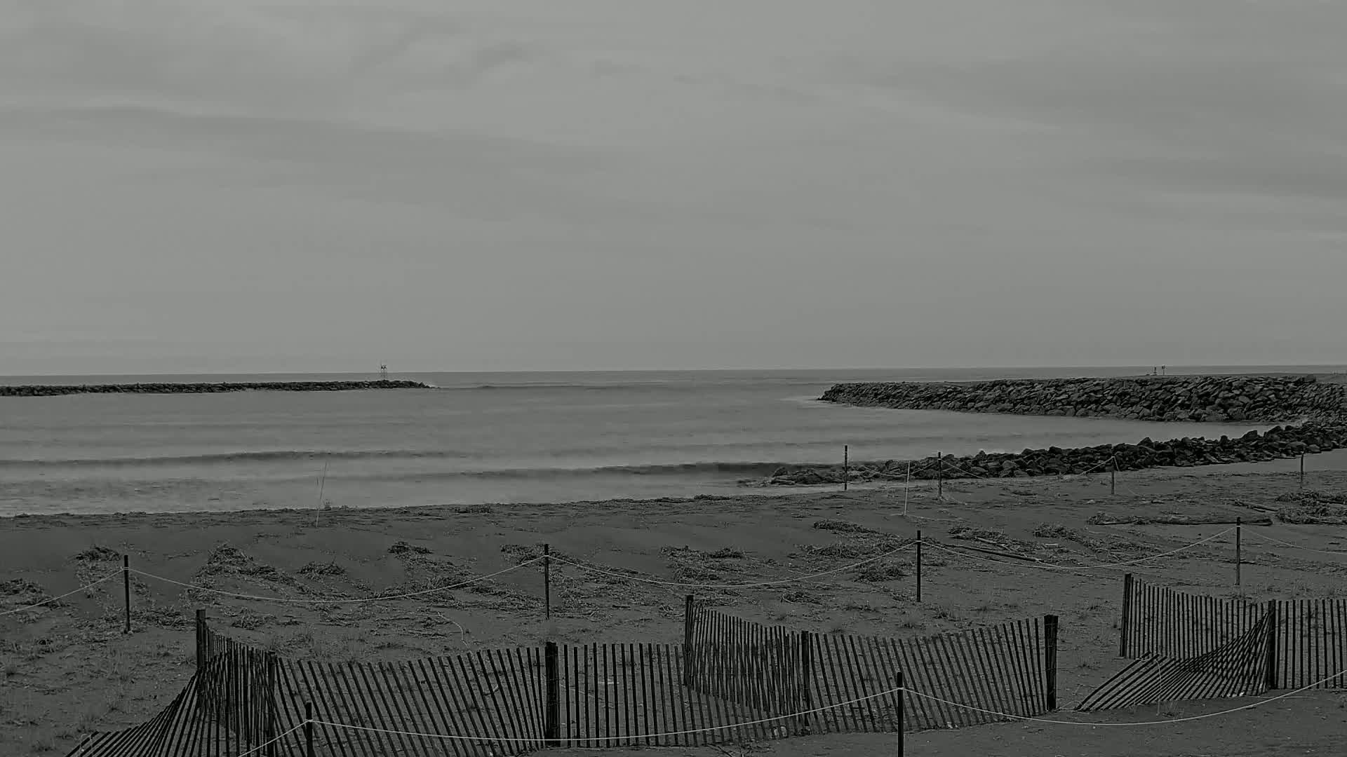 A gray, overcast sky hangs over a beach scene featuring gentle waves, rocky jetties, and several broken wooden fences.