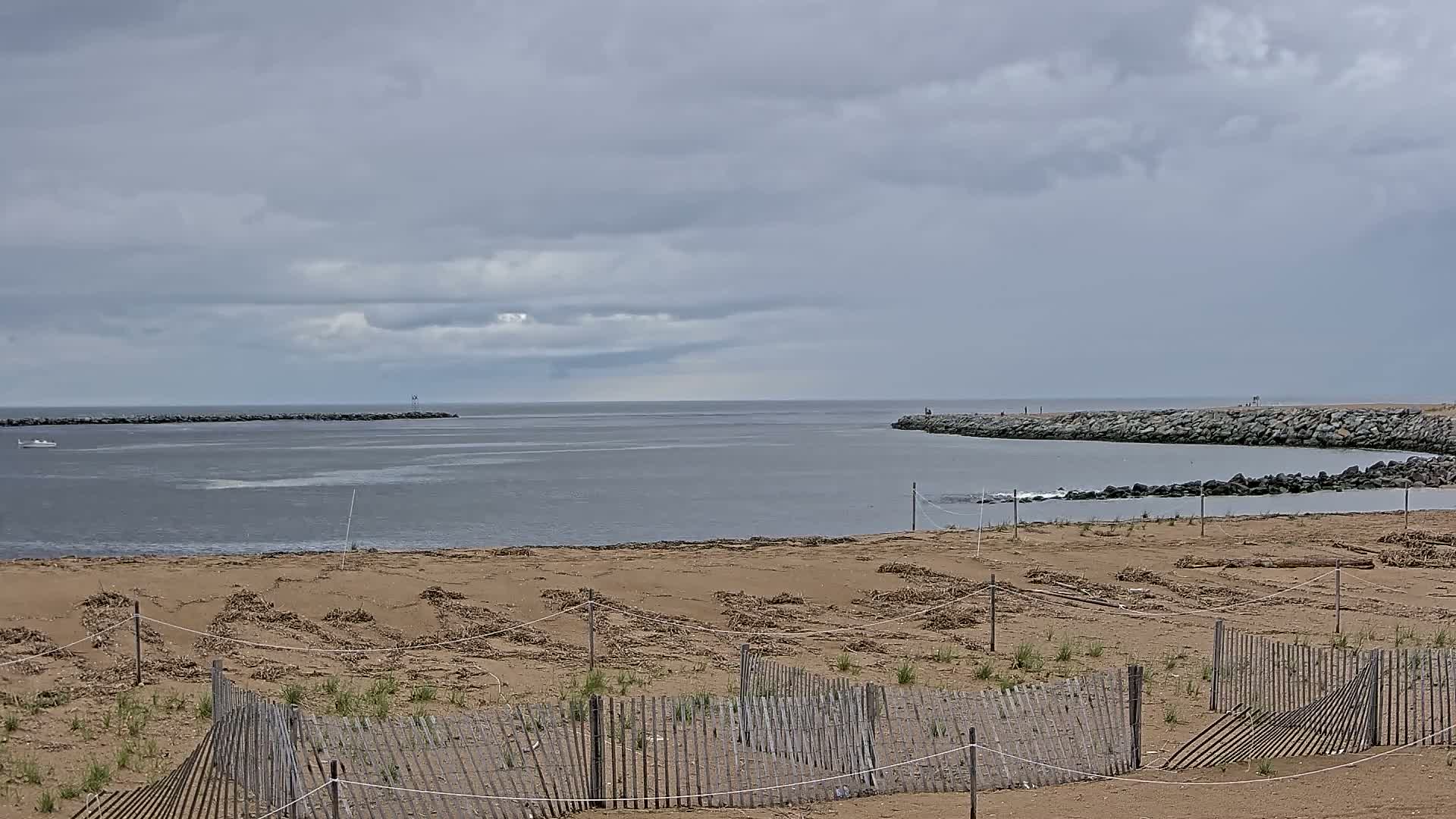 A sandy beach with low wooden fences stretches toward a calm bay under a cloudy sky, where a rocky jetty extends into the water.