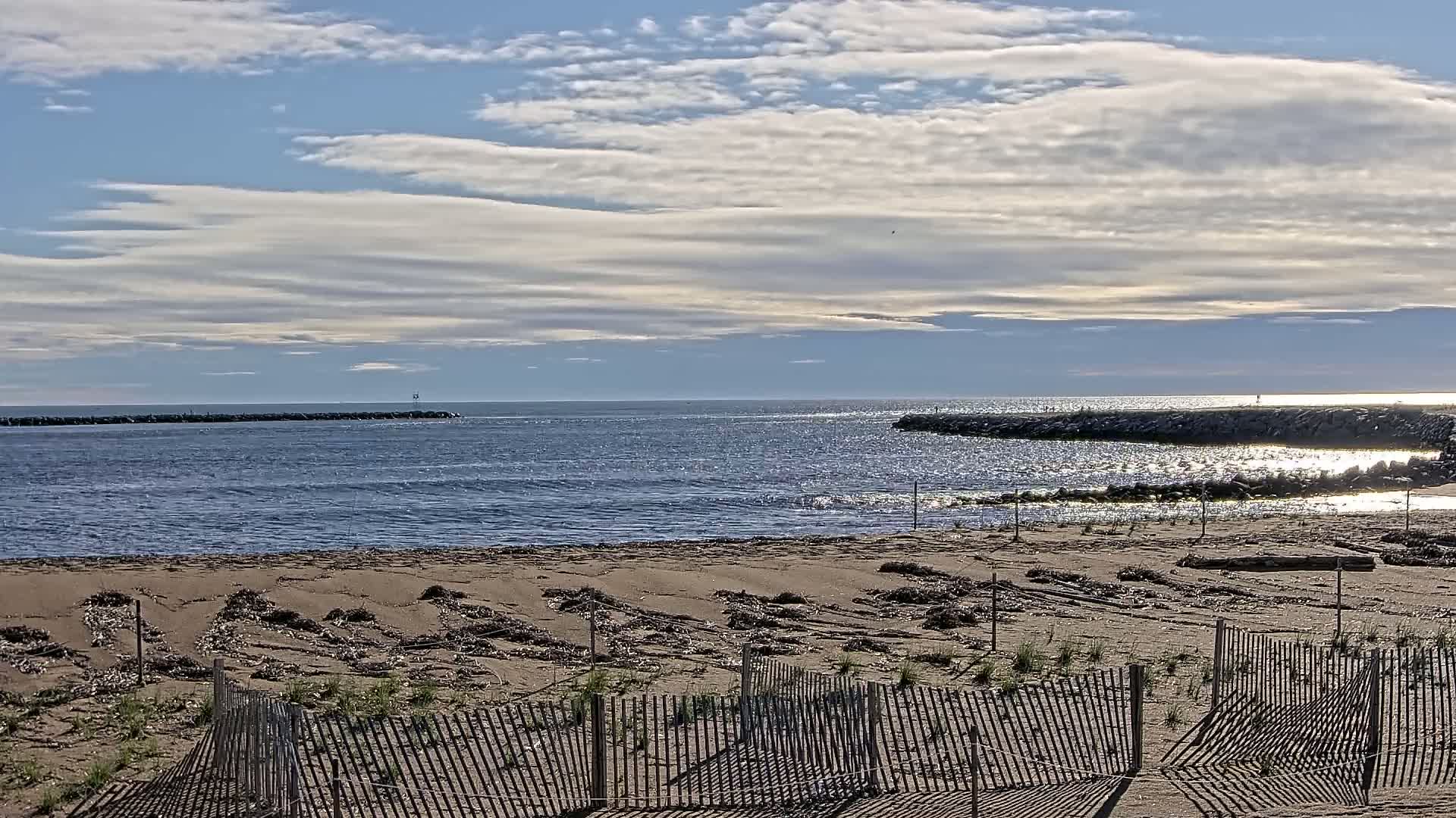 A sandy beach with low wooden fences stretches towards a calm ocean under a partly cloudy sky.