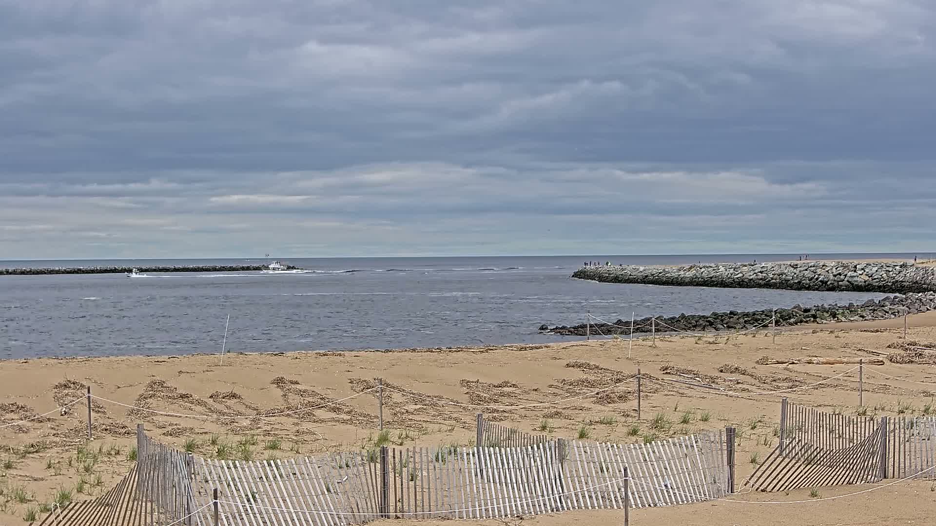 A sandy beach with a wooden fence is in the foreground, overlooking a calm inlet with a few boats and a rock jetty under a cloudy sky.