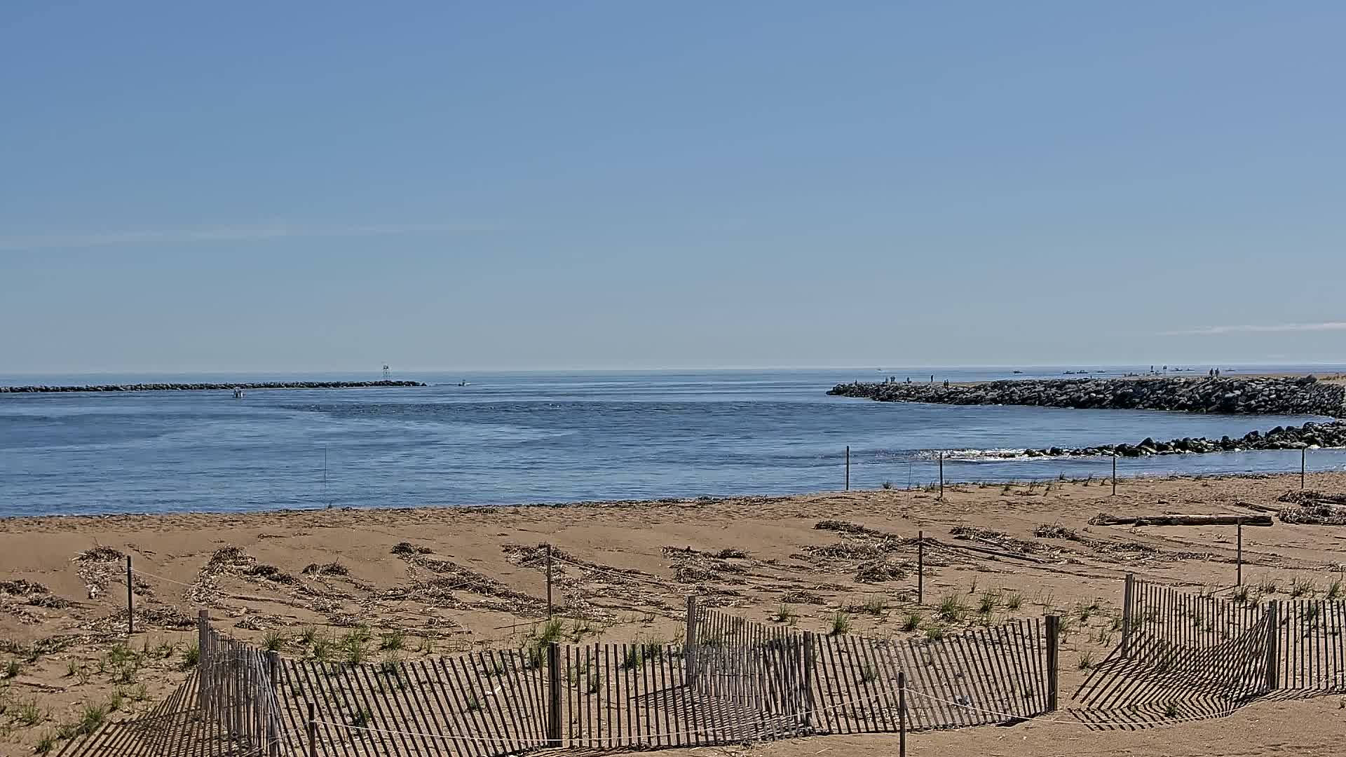 A sandy beach with a wooden fence stretches towards a calm ocean inlet, bordered by rock jetties under a clear blue sky.