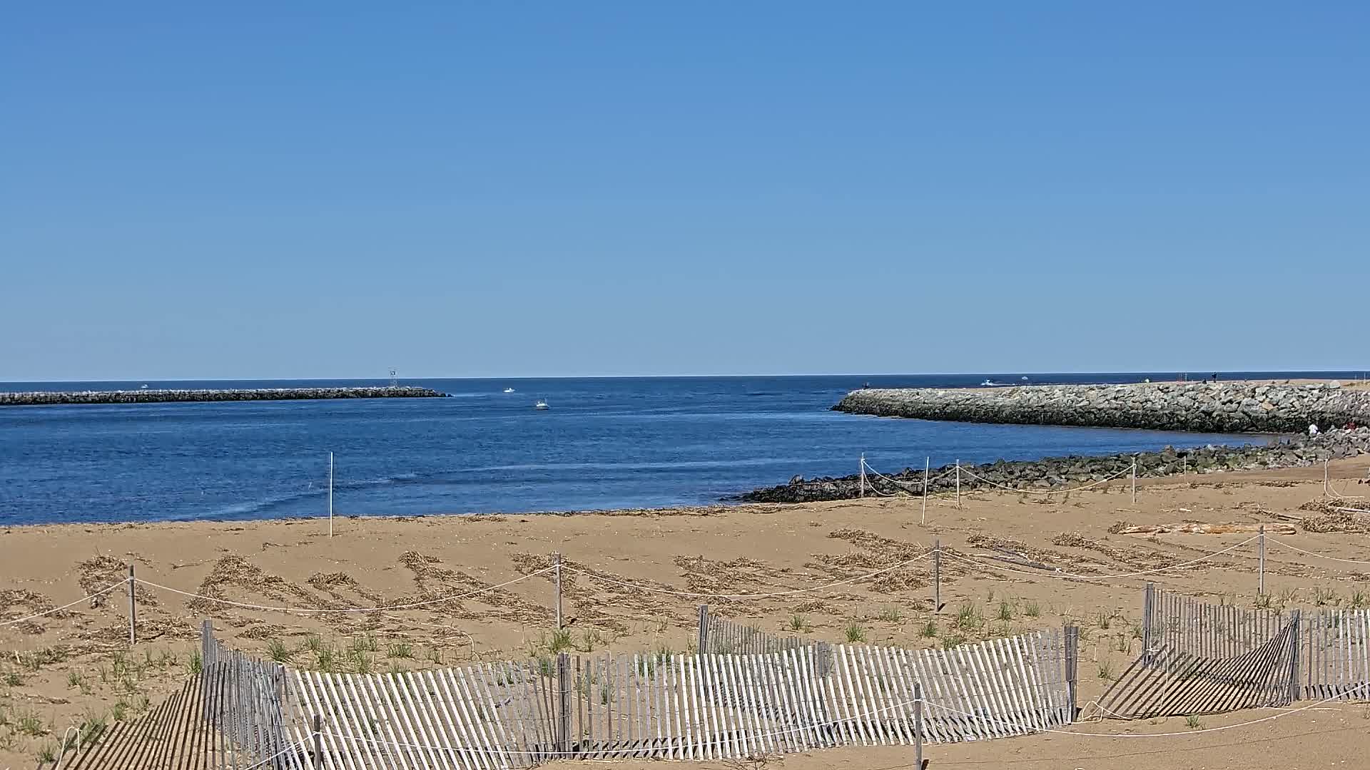 A sandy beach with low wooden fences stretches toward a calm, blue ocean inlet bordered by rock jetties under a clear blue sky.