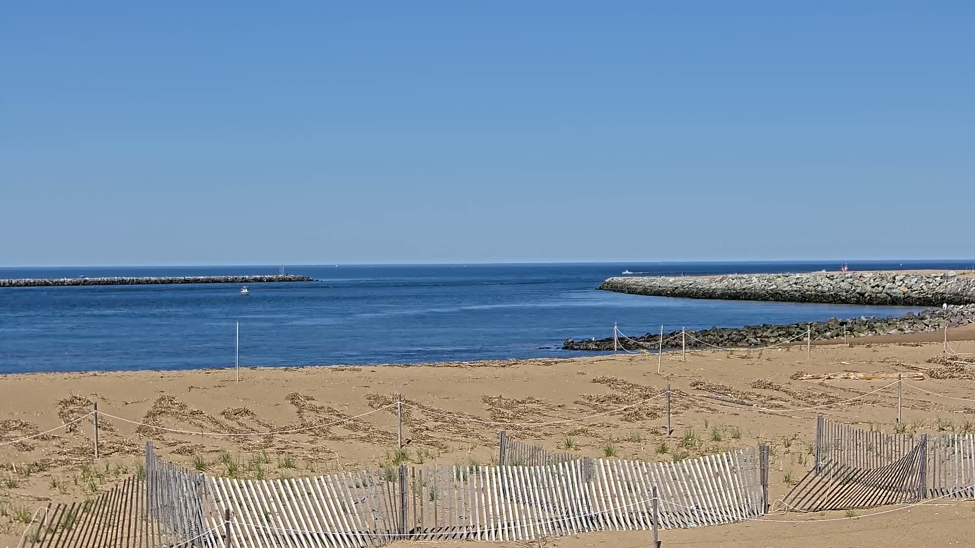 A sandy beach with low wooden fences and scattered seaweed lies before a calm blue ocean, partially enclosed by stone jetties on a clear, sunny day.