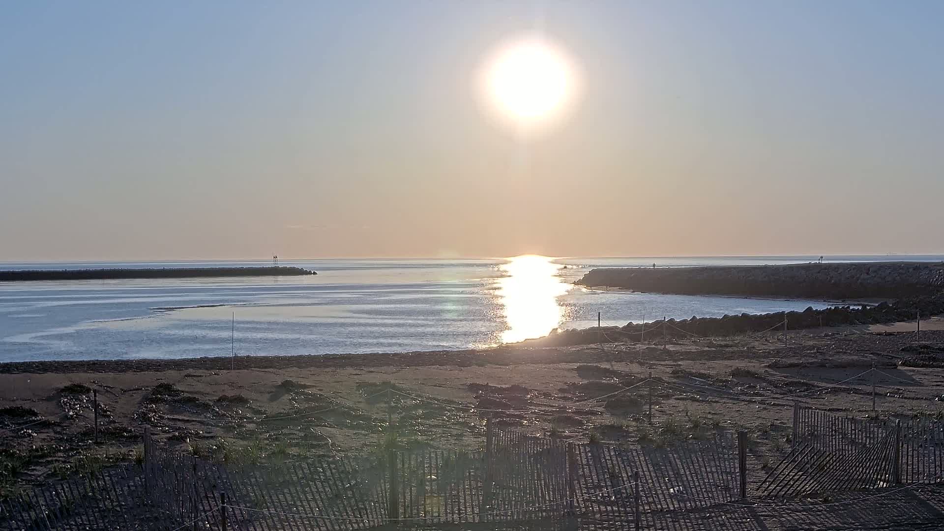 A sandy beach with low wooden fences is seen at sunrise, with calm water reflecting the bright sun and dark rock jetties extending into the ocean.