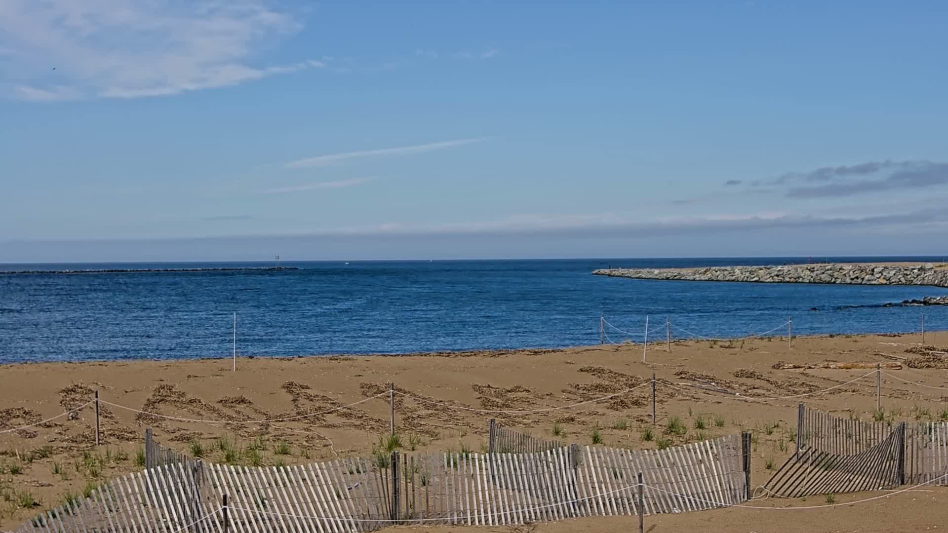 A sandy beach with a wooden fence in the foreground overlooks a calm, blue ocean under a partly cloudy sky.