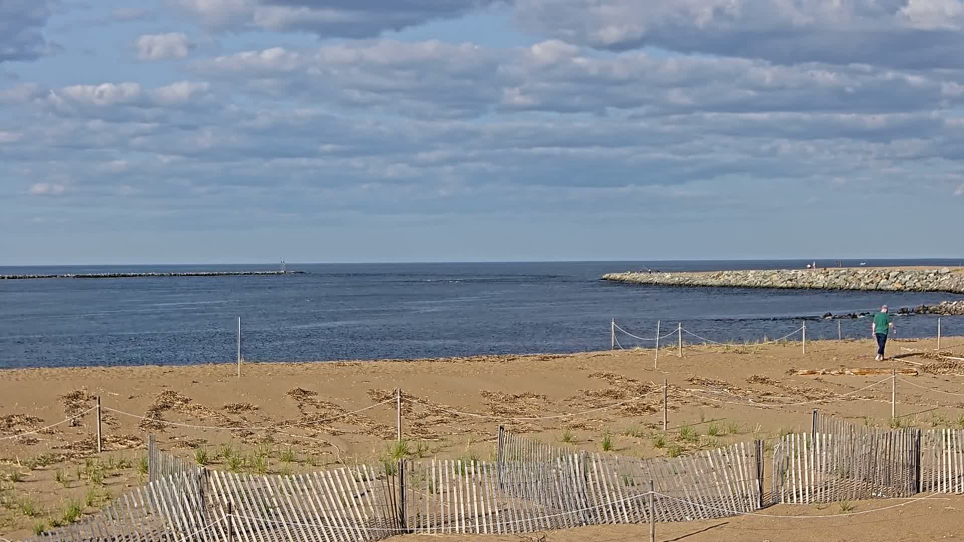 A sandy beach with a wooden fence and a person walking towards the water, under a partly cloudy sky.