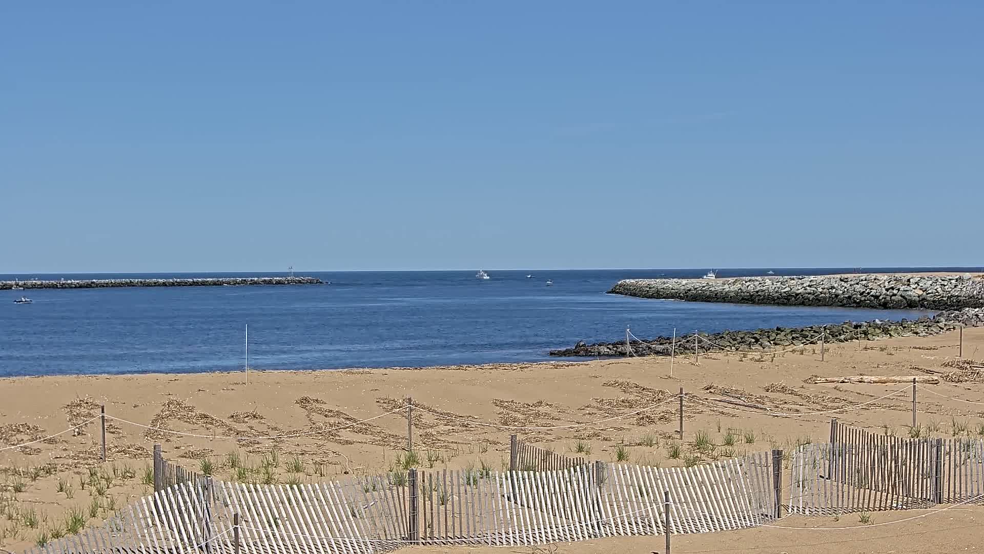 A sandy beach with a wooden fence is in the foreground, looking out to a calm ocean with several small boats and rock jetties under a clear blue sky.