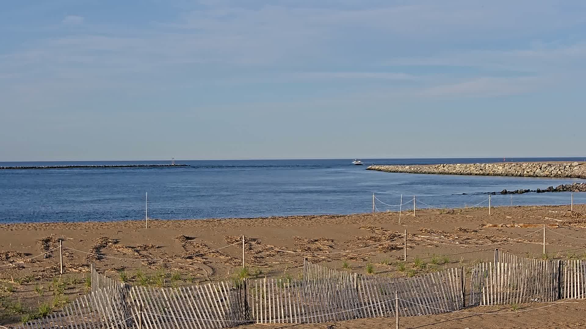 A sandy beach with a wooden fence and scattered seaweed lies before a calm inlet of blue water, bordered by rock jetties under a clear, bright sky.