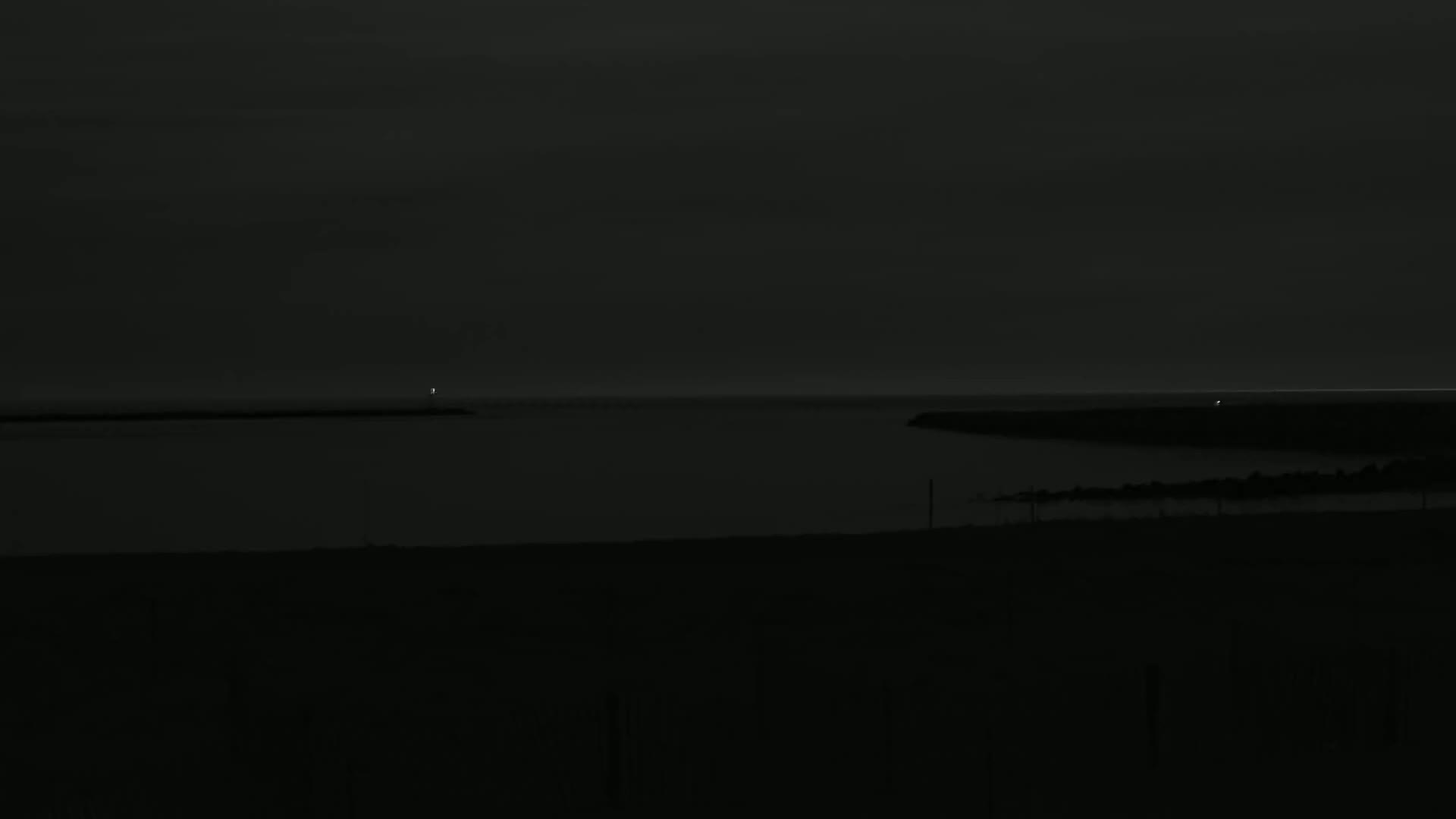 A dark, nighttime scene shows a calm body of water with a faint light in the distance, under a mostly clear sky.