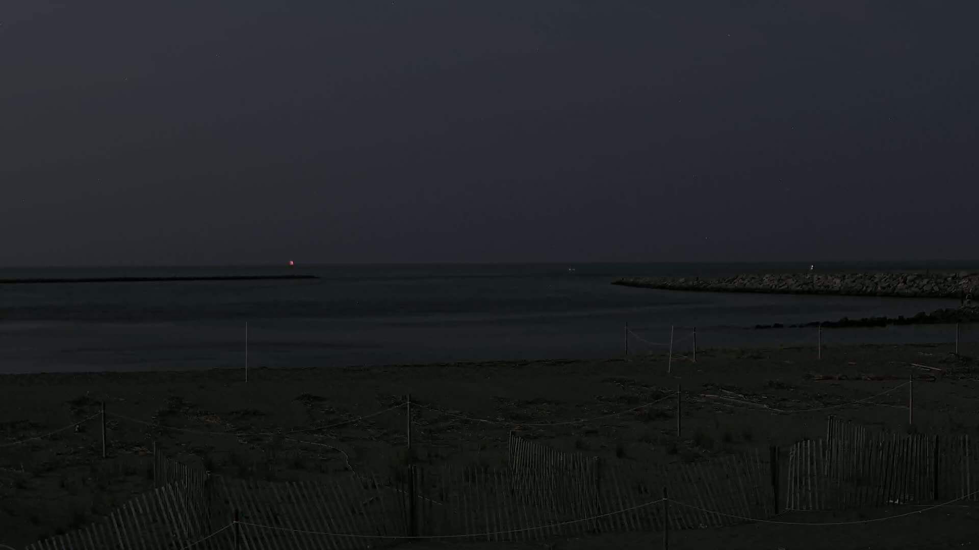 A dark beach at night with a calm, dark body of water beyond a low rock jetty, under a mostly clear, dark sky.