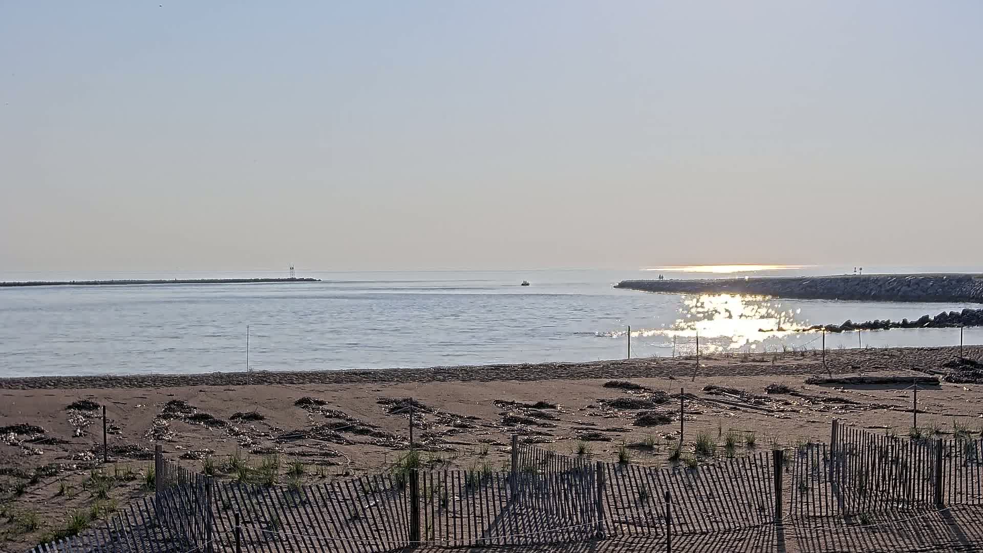 A sandy beach with a wooden fence in the foreground overlooks calm water reflecting sunlight, with two long jetties extending into the distance under a clear sky.
