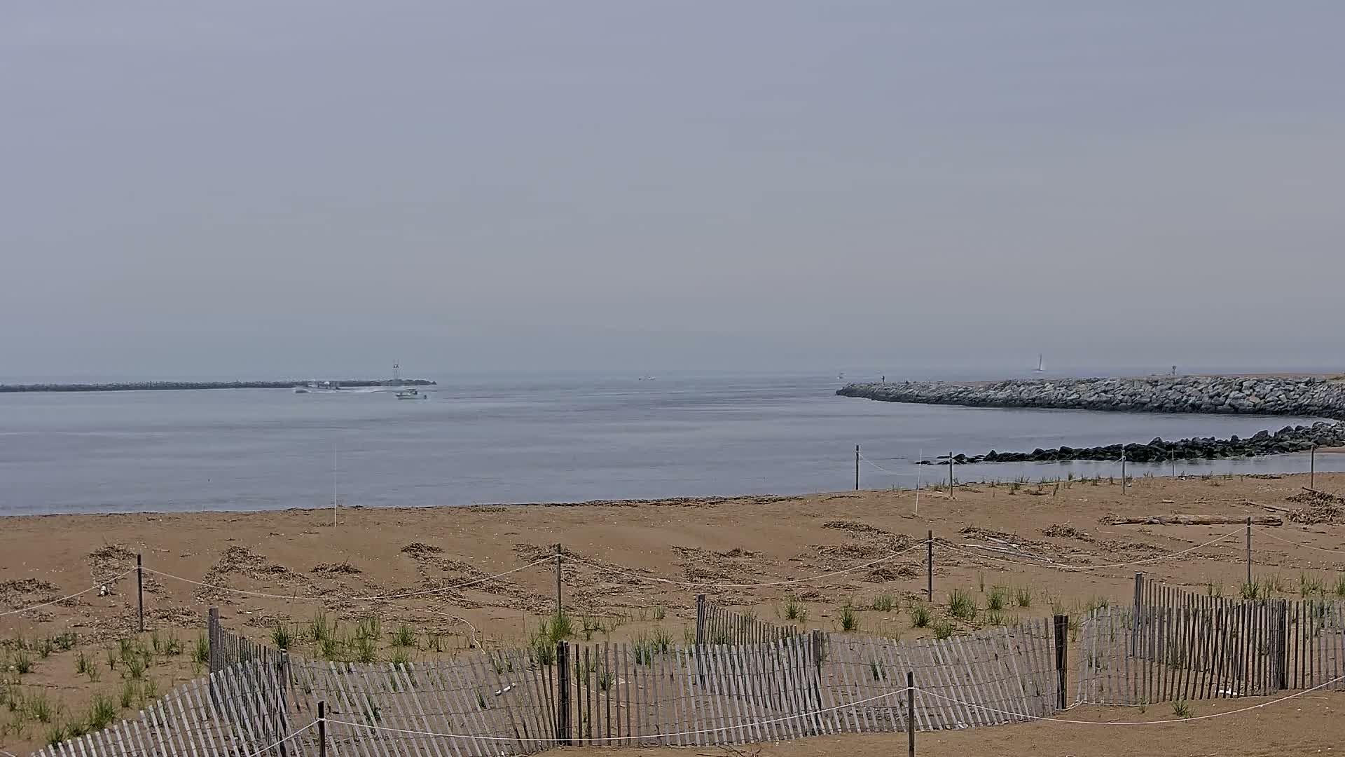 A sandy beach with a wooden fence in the foreground overlooks calm water and rock jetties on a hazy day.