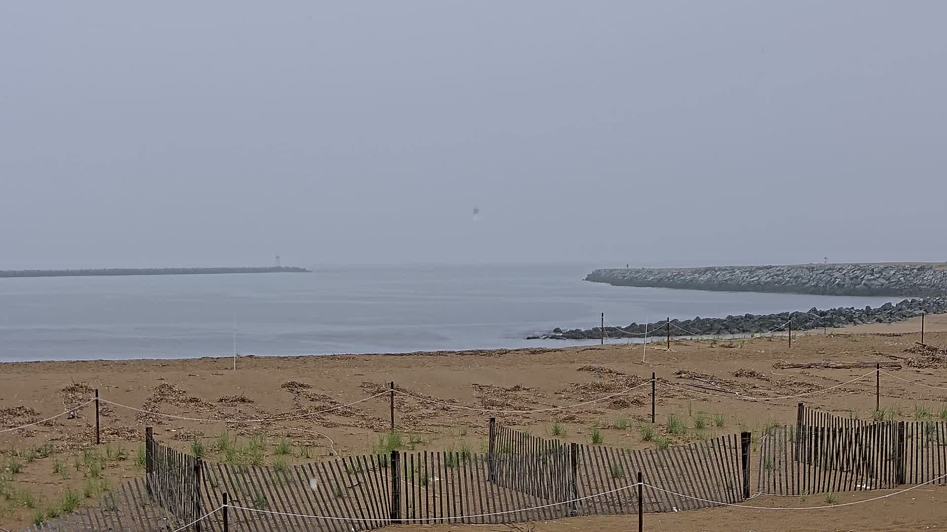 A sandy beach with low wooden fences protecting young vegetation is visible, overlooking calm, foggy water and distant rock jetties.
