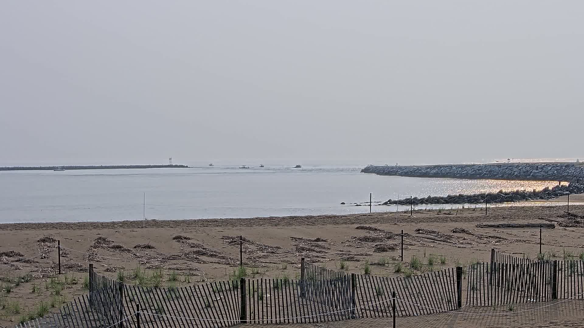 A hazy beach scene shows a calm body of water with several small boats, separated from a sandy beach by a low-lying jetty, all protected by a simple wooden fence.