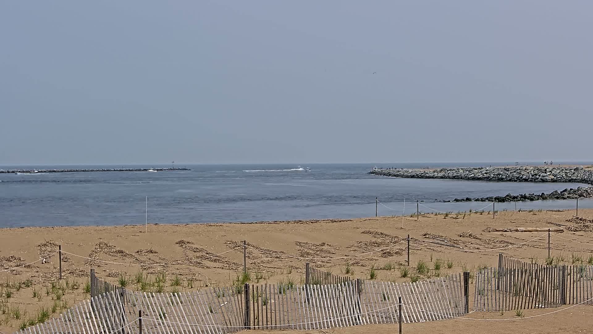 A sandy beach with a wooden fence is shown, overlooking a calm body of water with jetties on either side under a hazy sky.