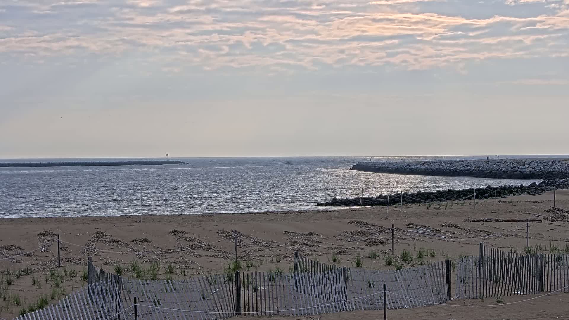 A sandy beach with a low wooden fence stretches towards a calm body of water, partially enclosed by rock jetties under a cloudy sky.