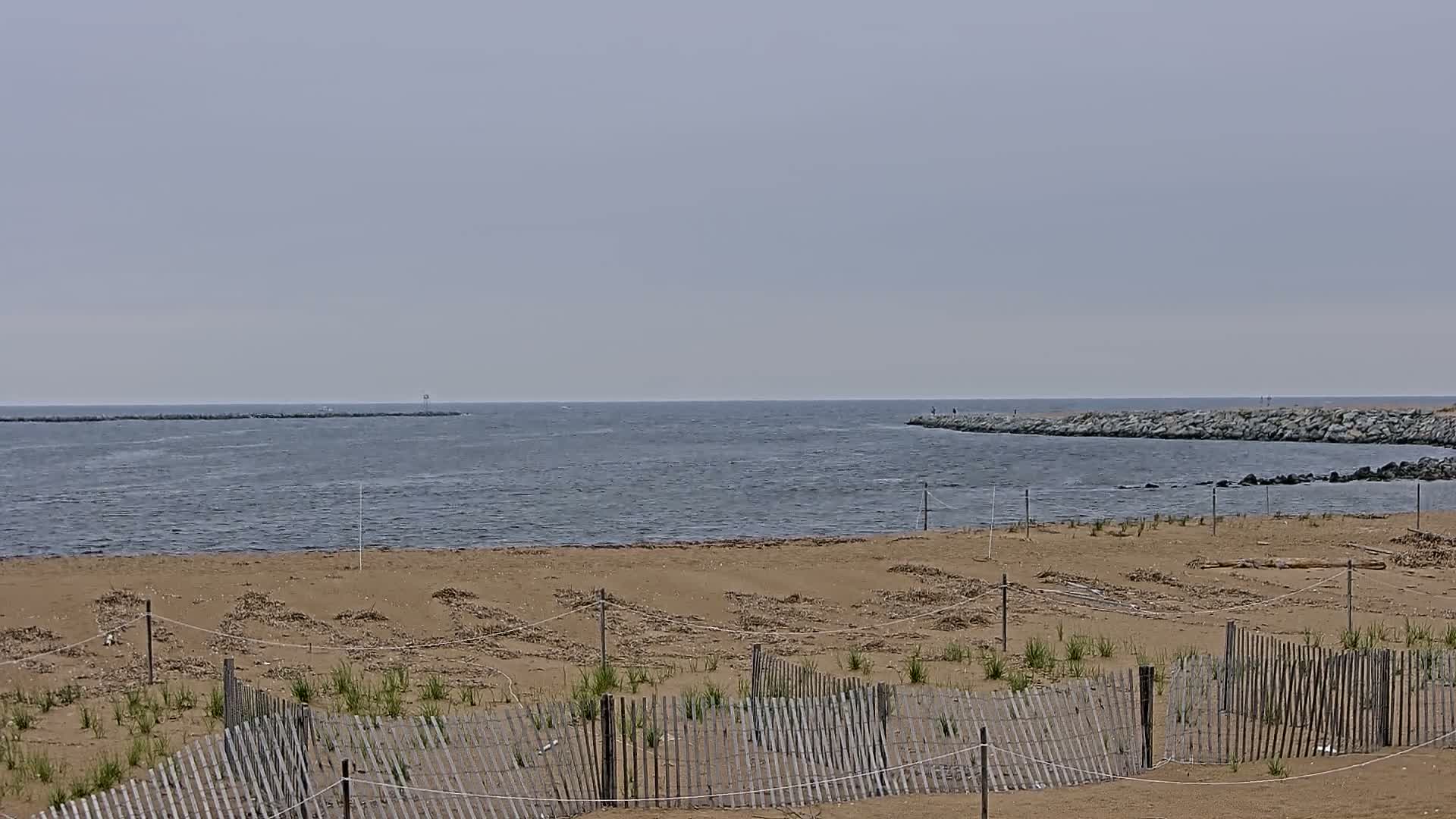 A sandy beach with a wooden fence and young vegetation is visible, overlooking a calm body of water with rock jetties on either side under a hazy sky.