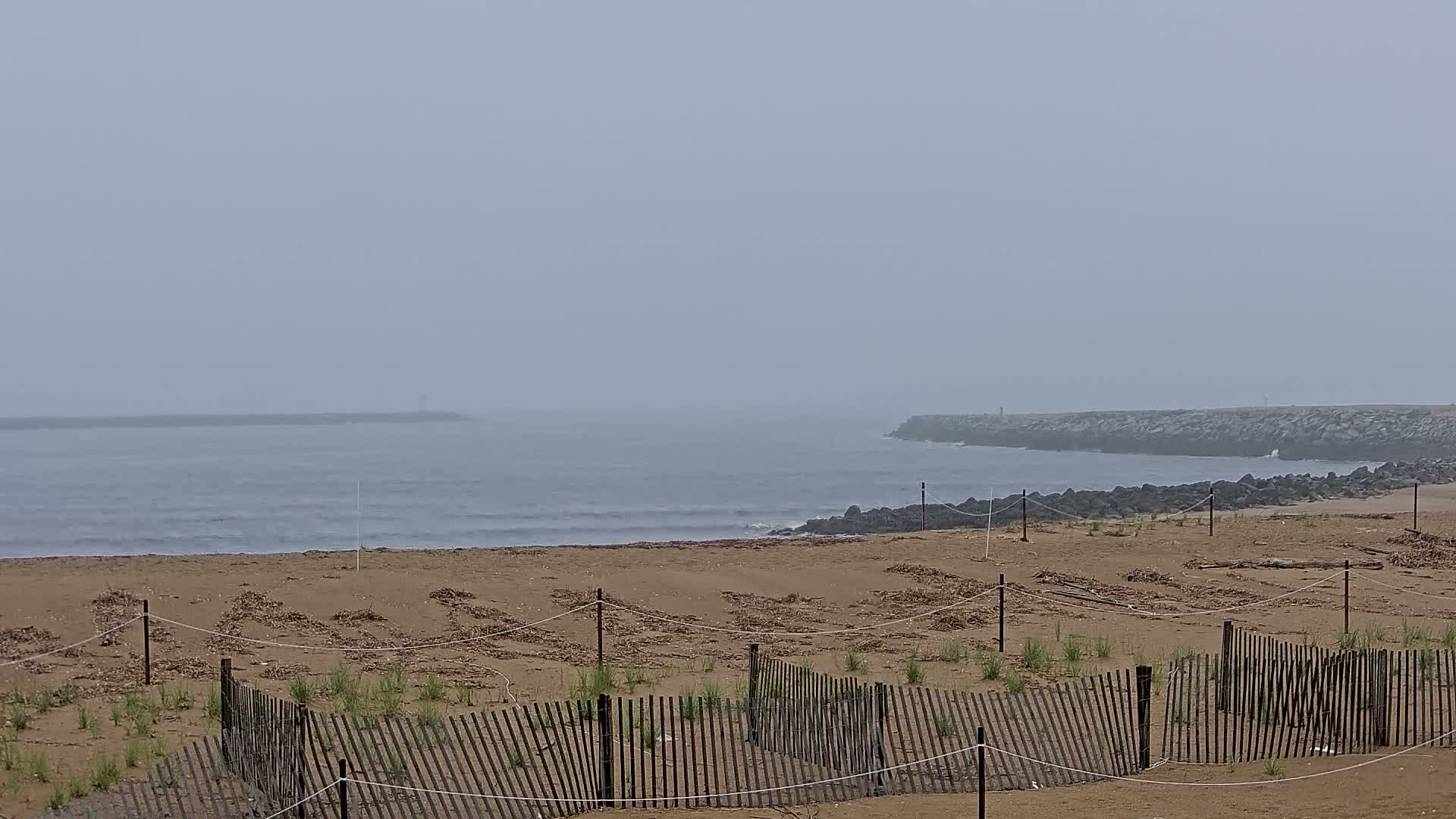 A foggy beach scene shows a sandy shore with short wooden fences protecting sparse vegetation, and a rocky jetty in the distance.