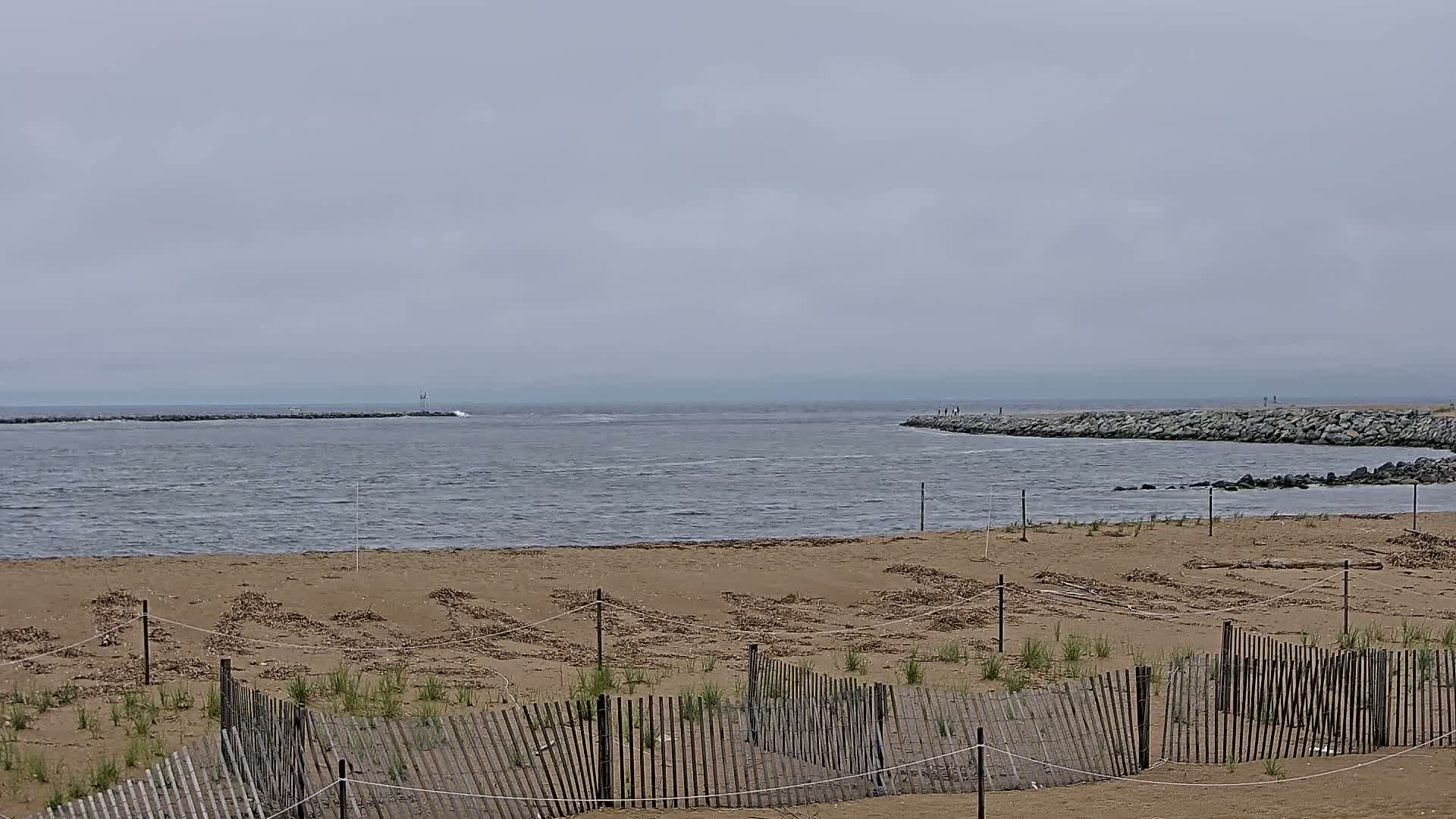 A sandy beach with low wooden fences is seen on an overcast day, overlooking a calm, grey body of water with rock jetties extending into it.