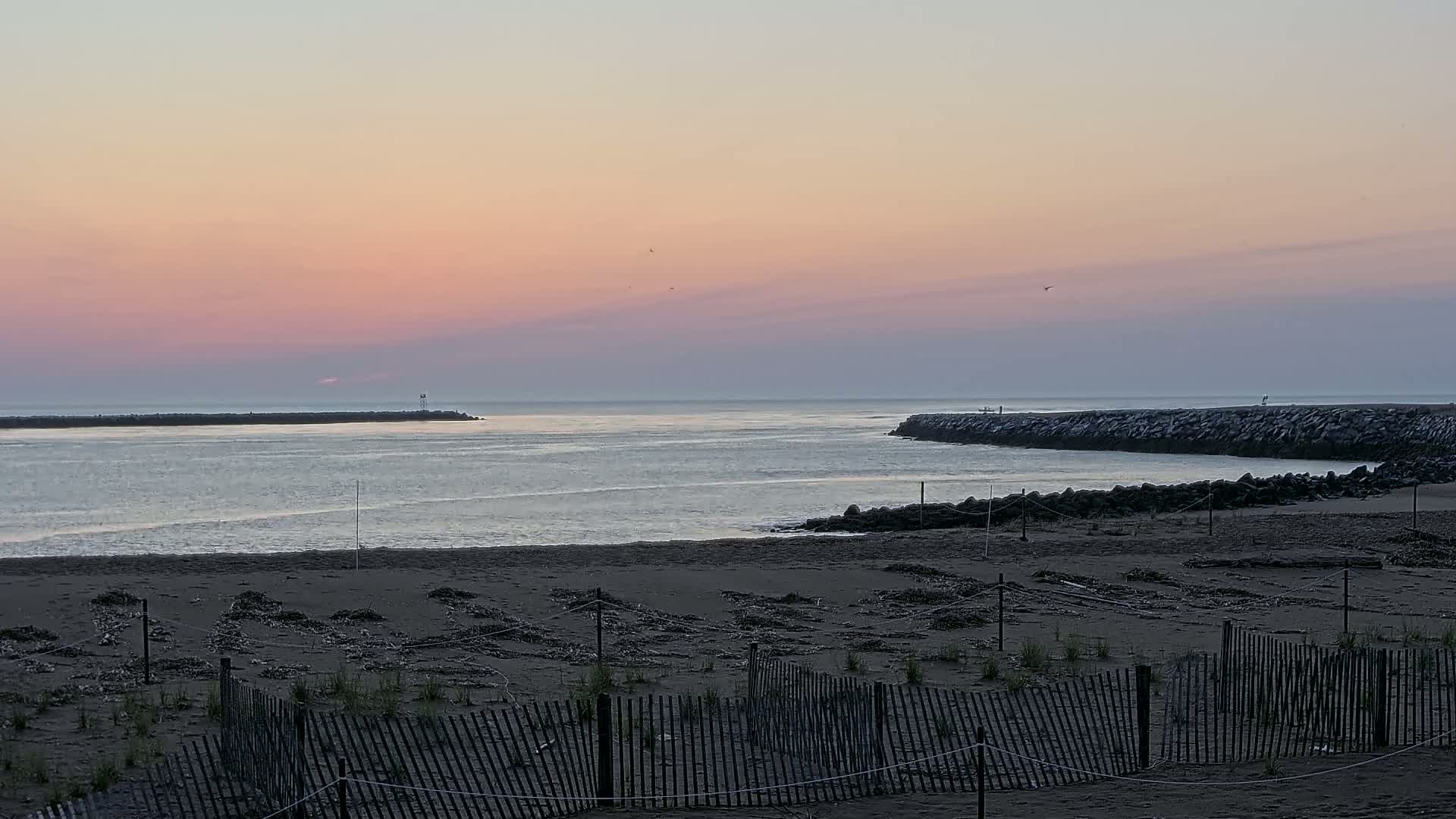 A calm ocean inlet at sunrise, viewed from a sandy beach behind a low wooden fence.
