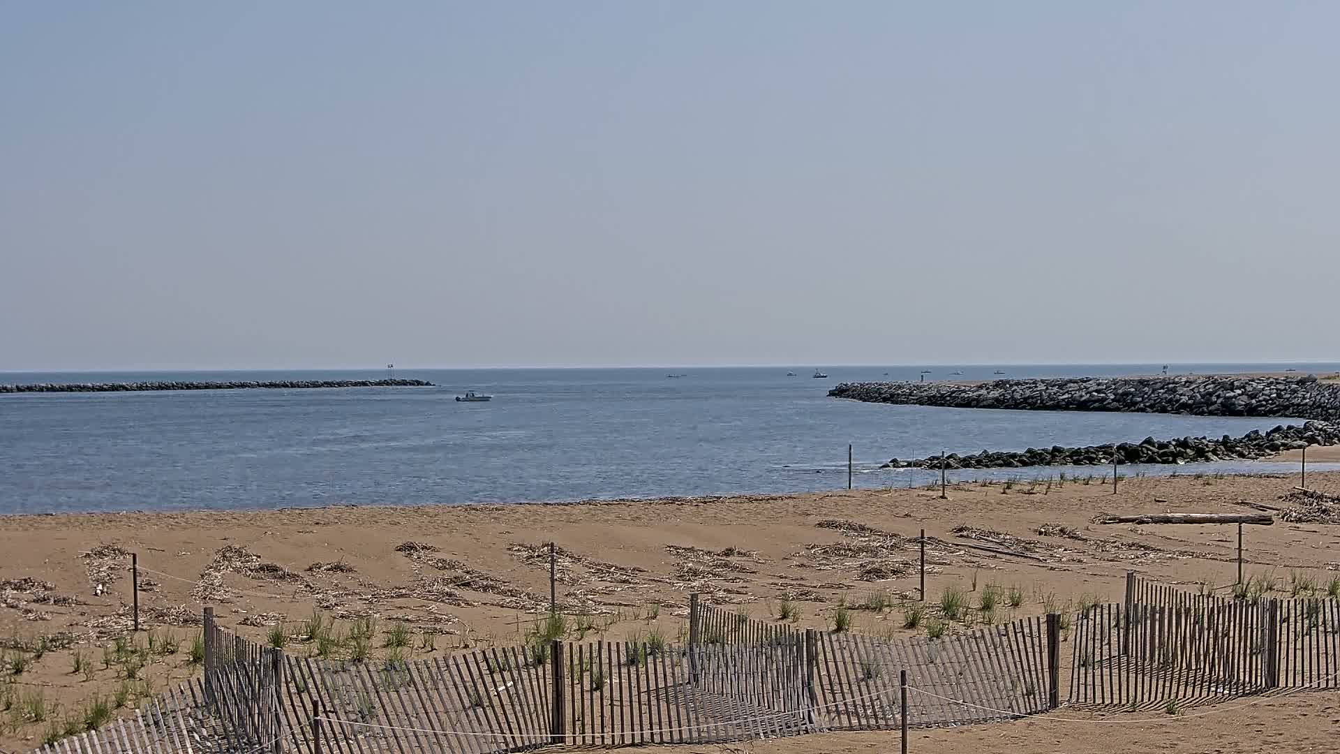 A sandy beach with a wooden fence in the foreground overlooks calm water with a few small boats and rock jetties on a clear, sunny day.