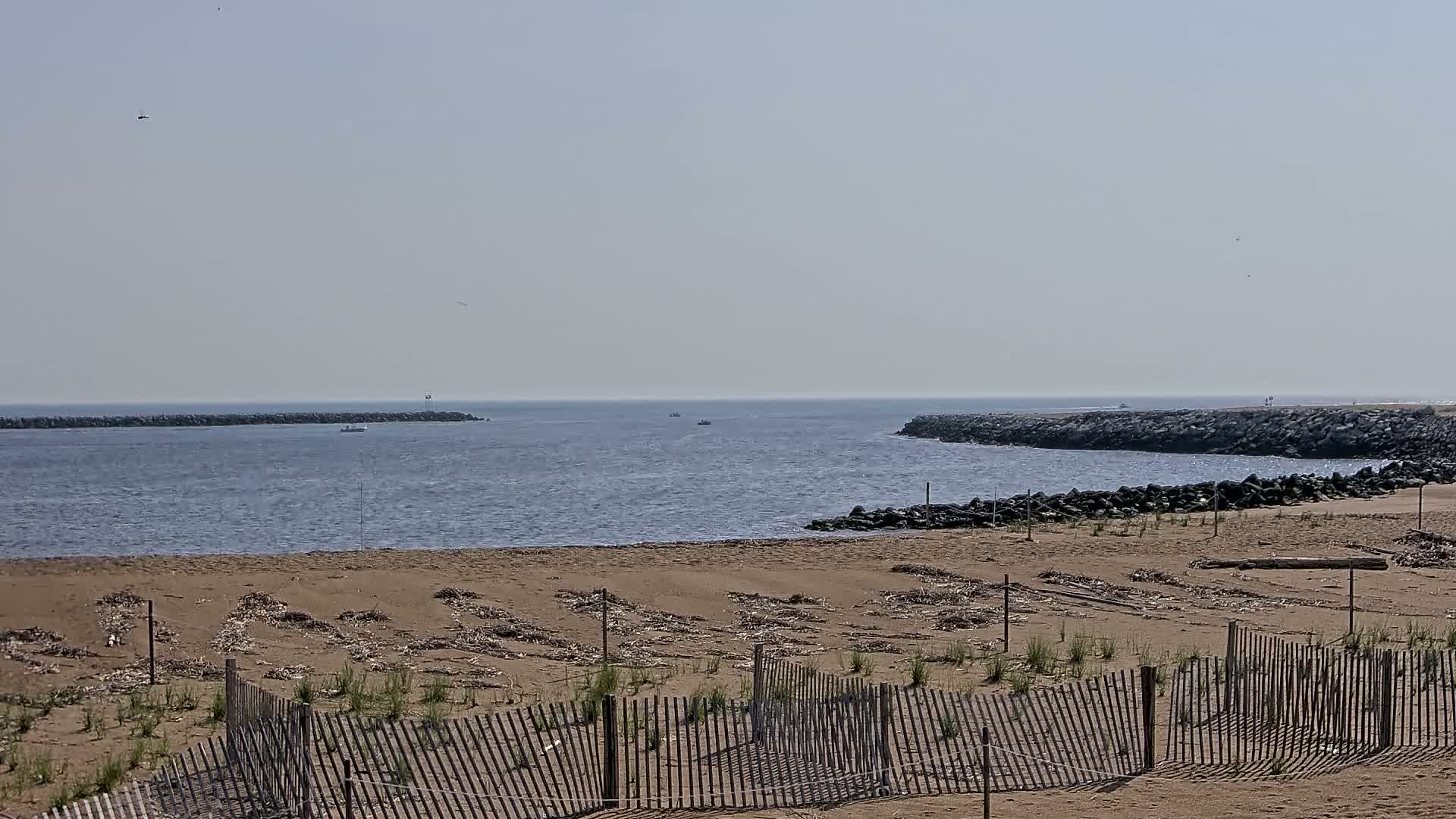 A sandy beach with a wooden fence is seen in the foreground, overlooking calm ocean water with a few small boats and rock jetties on either side of the inlet under a clear, bright sky.