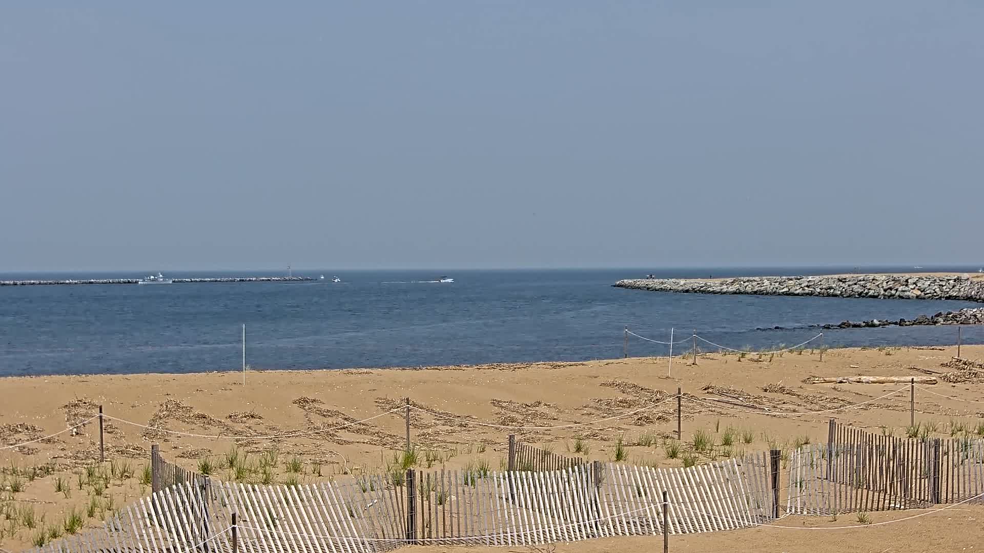 A sandy beach with a wooden fence, small plants, and tire tracks in the foreground overlooks calm blue water, a rock jetty, and several small boats under a clear sky.