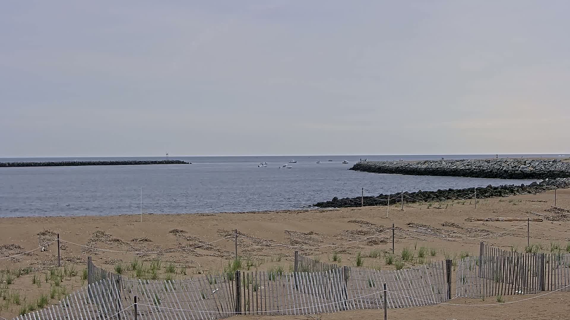 A sandy beach with a wooden fence is visible in the foreground, overlooking a calm body of water with several small boats and rock jetties under an overcast sky.
