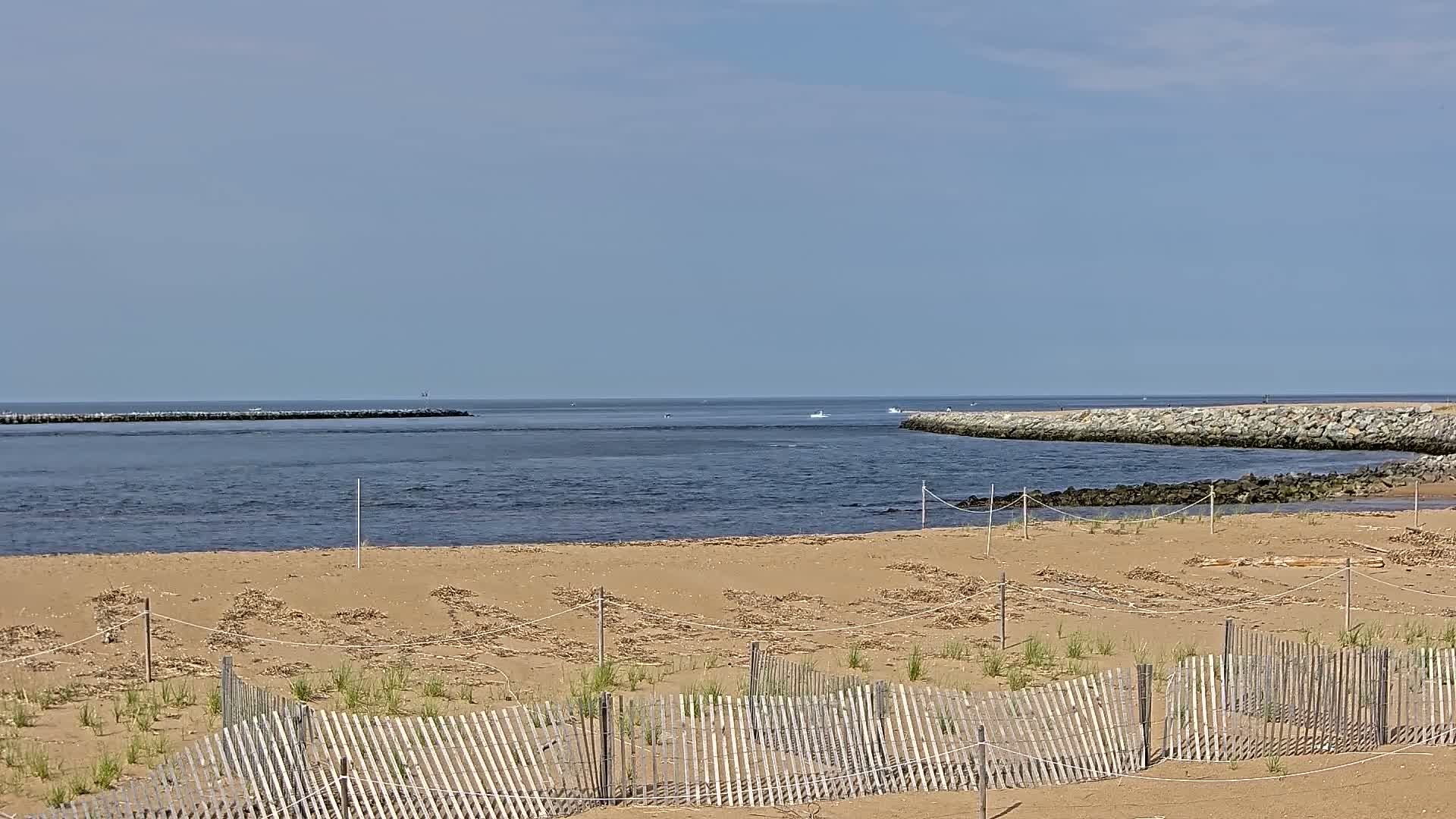 A sandy beach with a wooden fence and small plants, leading to a calm body of water with stone jetties on either side under a clear sky.