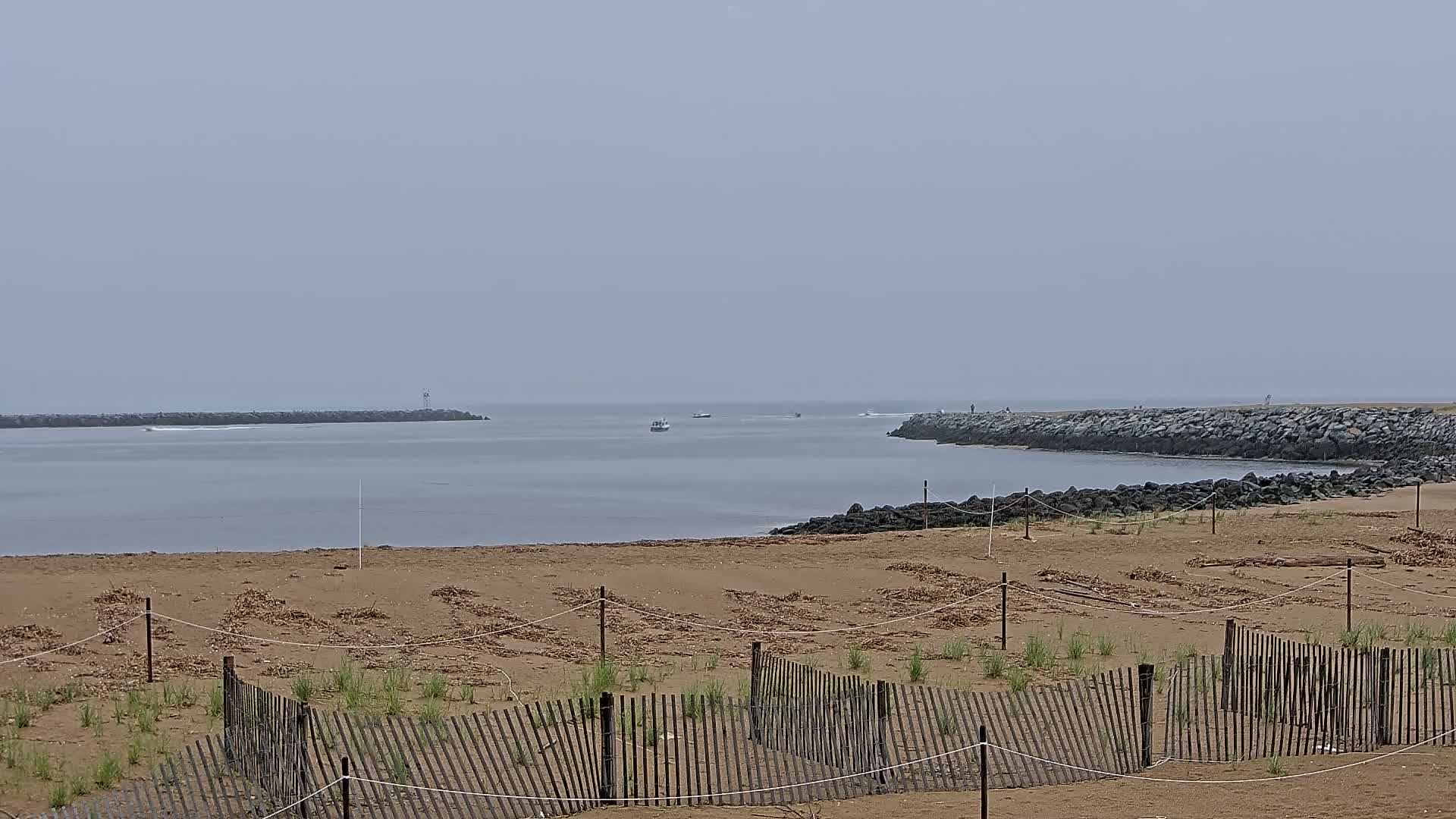 A sandy beach with a low wooden fence in the foreground overlooks a calm, gray body of water enclosed by rock jetties under an overcast sky.
