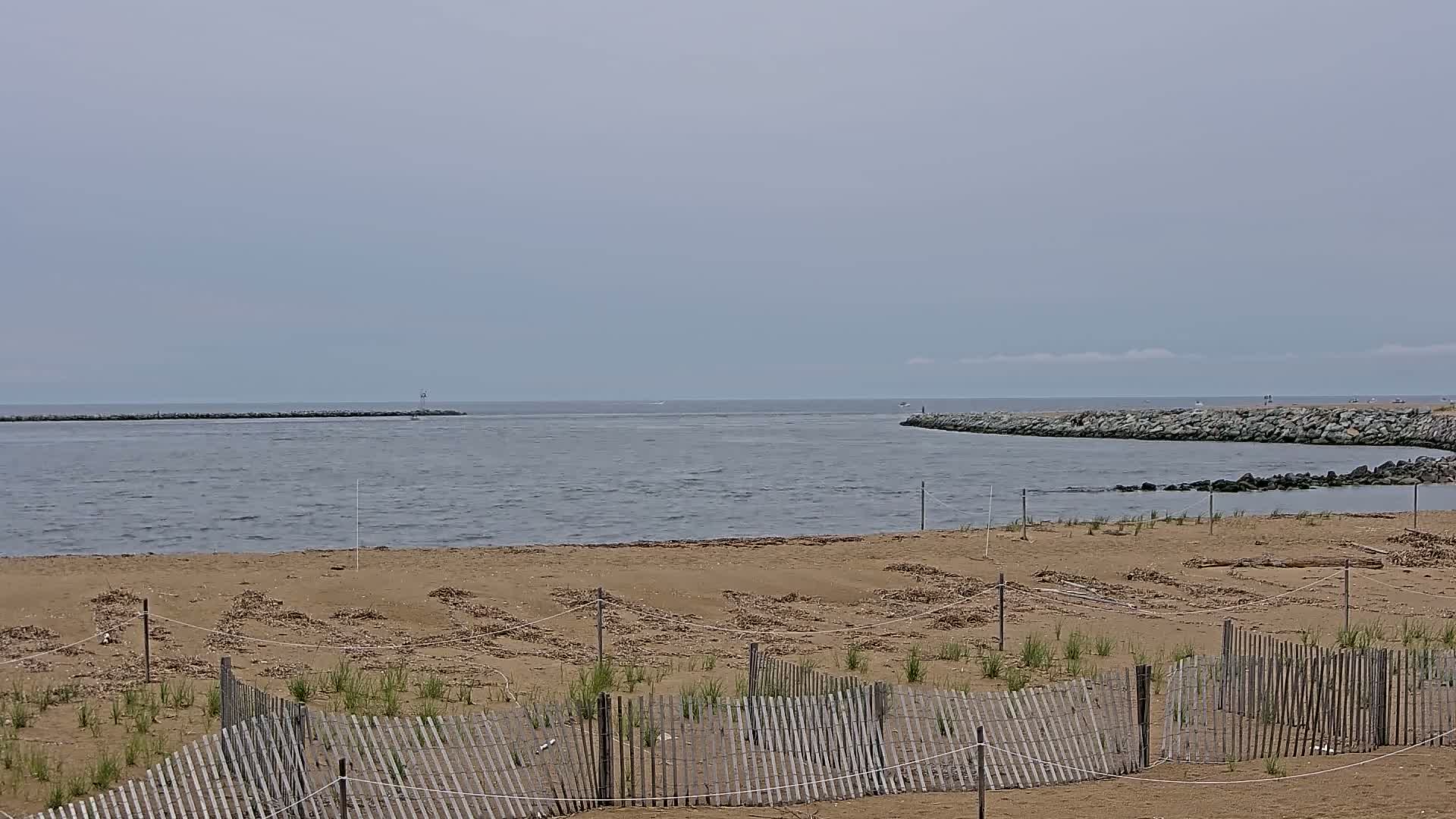 A sandy beach with a wooden fence and small plants is visible in the foreground, overlooking a calm, gray body of water with rock jetties on either side under an overcast sky.