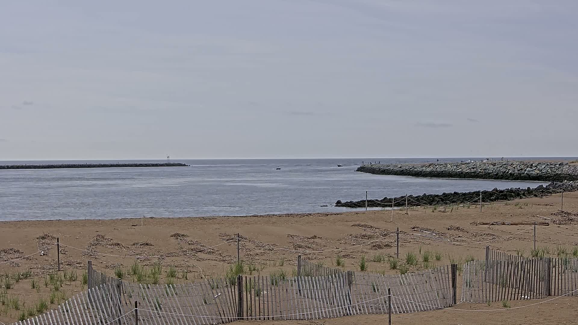 A sandy beach with a wooden fence and small plants is visible in the foreground, overlooking a calm, gray body of water with rock jetties on either side under an overcast sky.