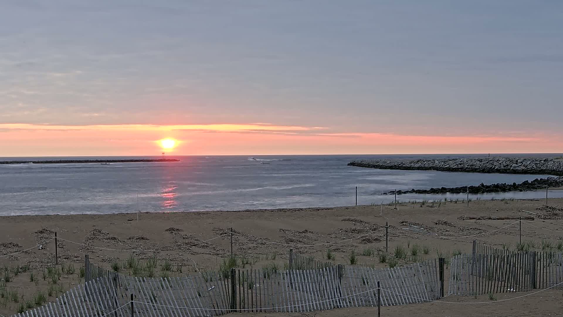 A sandy beach with a wooden fence overlooks calm ocean water at sunrise, under a pale pink and gray sky.