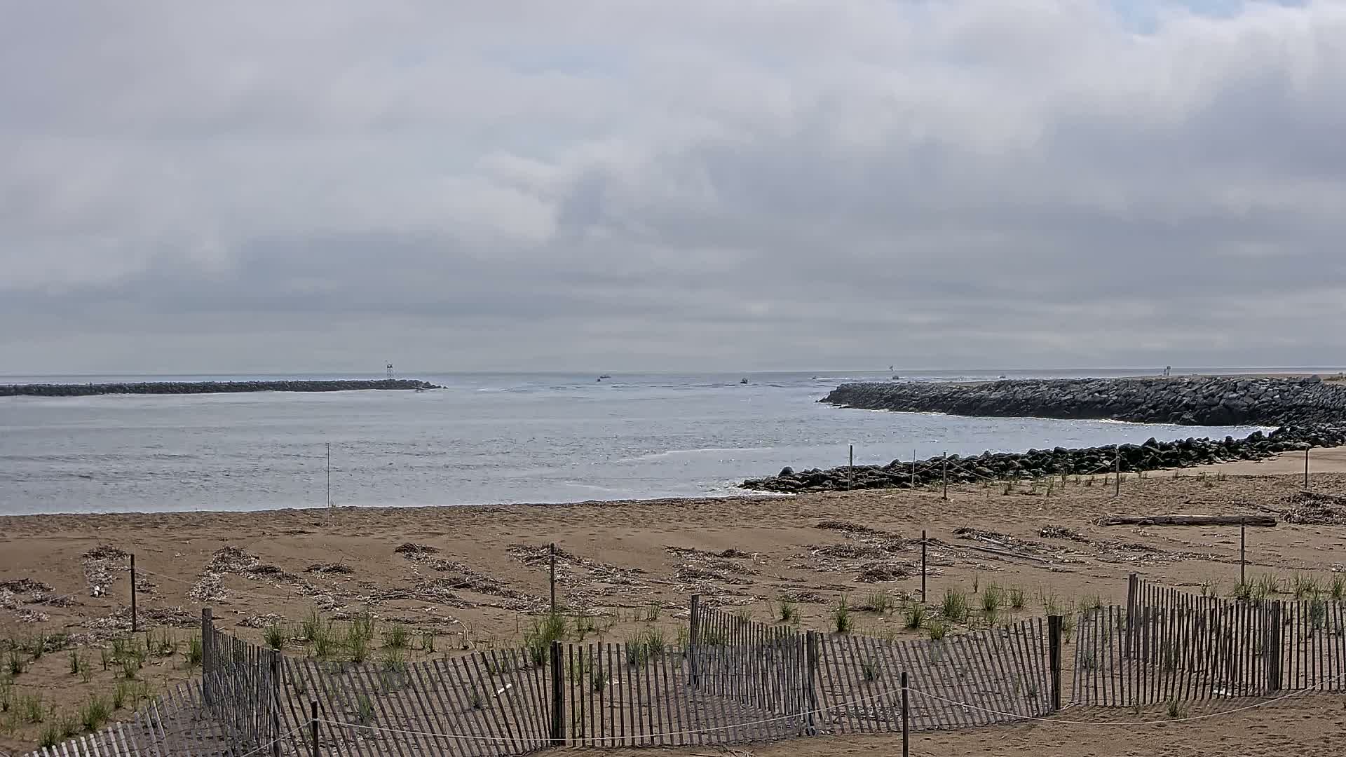 A sandy beach with a wooden fence is situated before a calm, gray-sky ocean inlet, which is bordered by dark rock jetties.