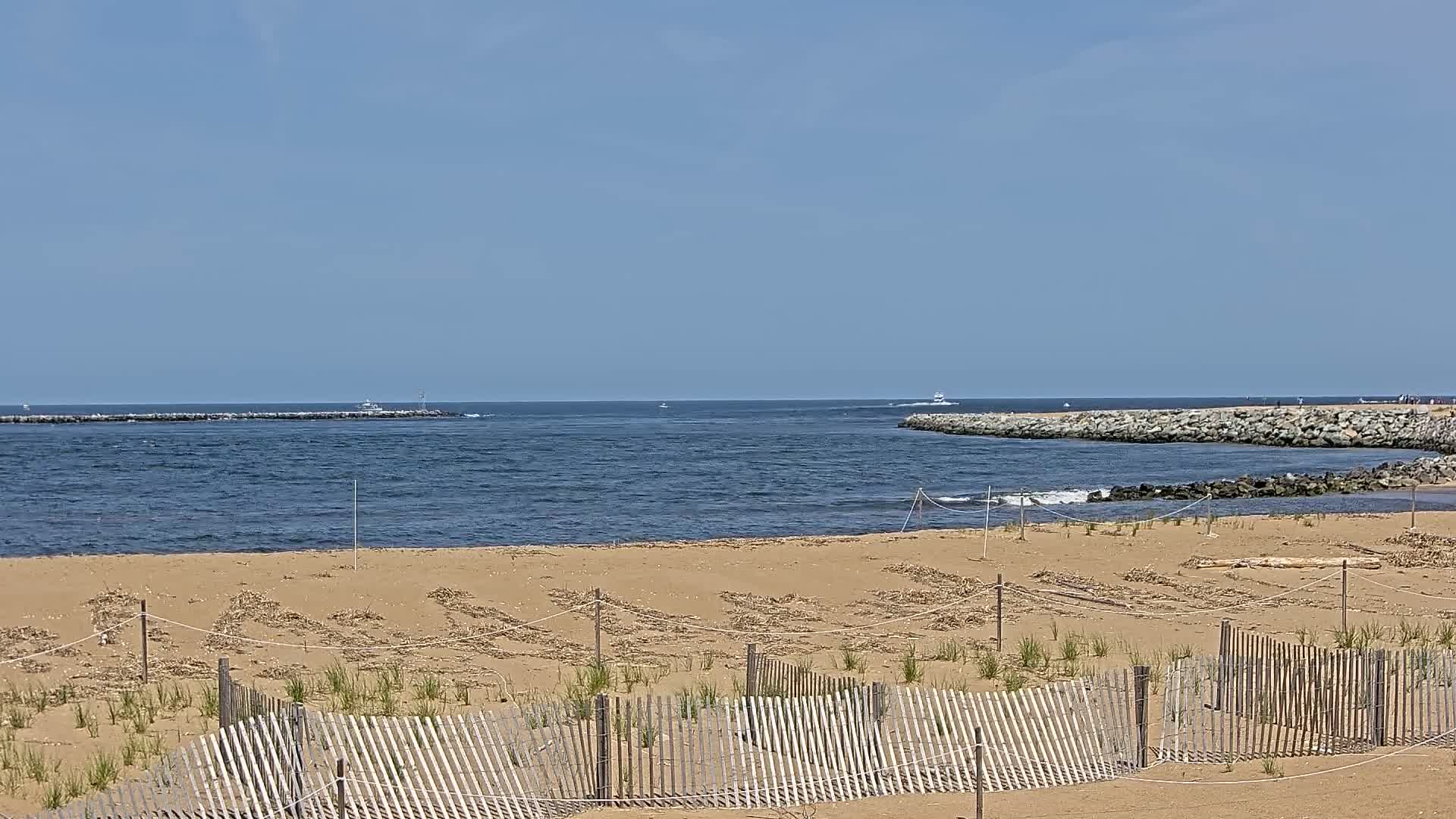 A sandy beach with small wooden fences and sparse vegetation overlooks a calm ocean inlet under a clear blue sky.