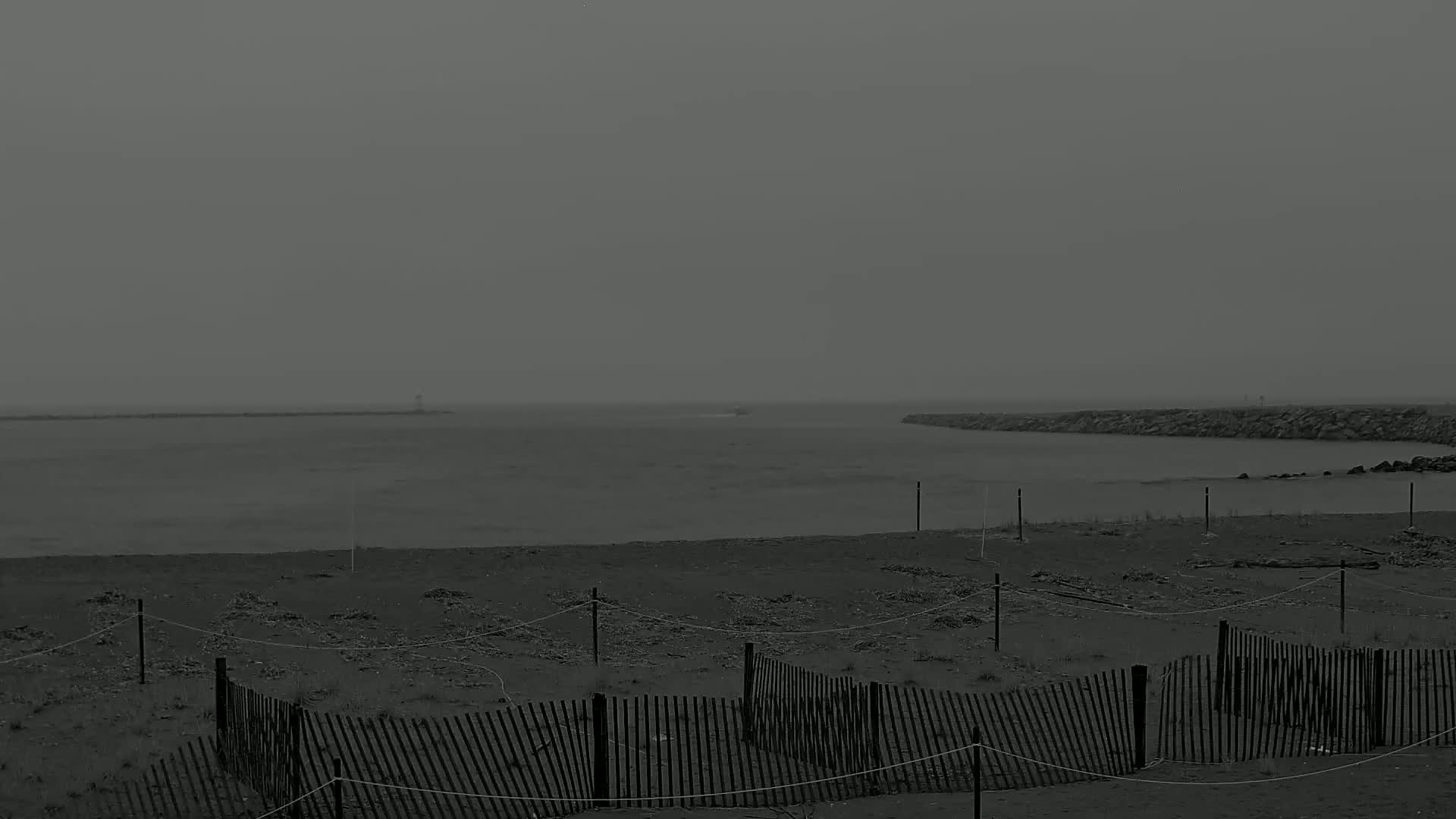 A gray, overcast day shows a beach with short, broken wooden fences, leading to a calm body of water with a breakwater in the distance.