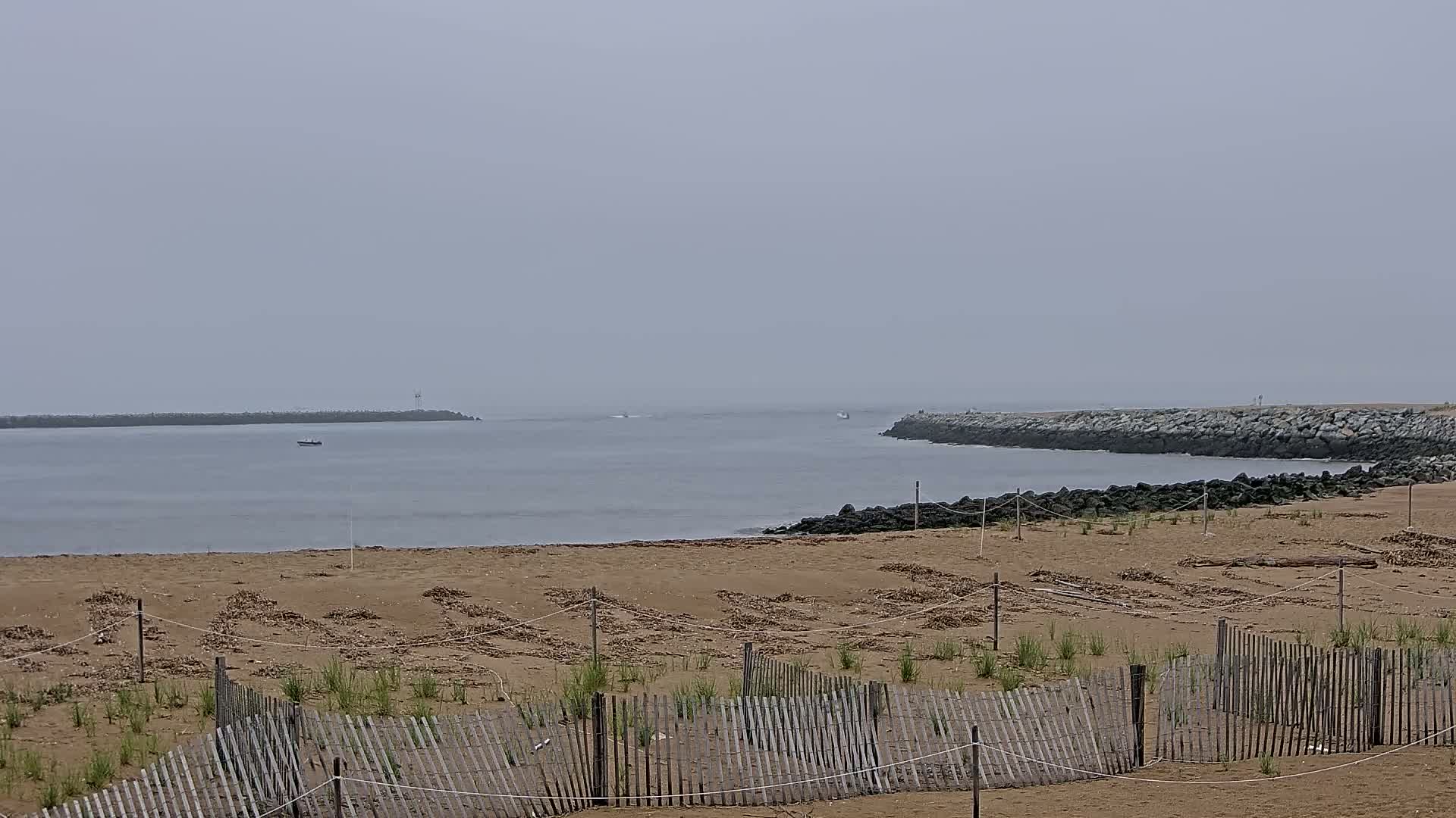 A sandy beach with a wooden fence and small plants stretches toward a calm, gray body of water partially enclosed by rock jetties under an overcast sky.