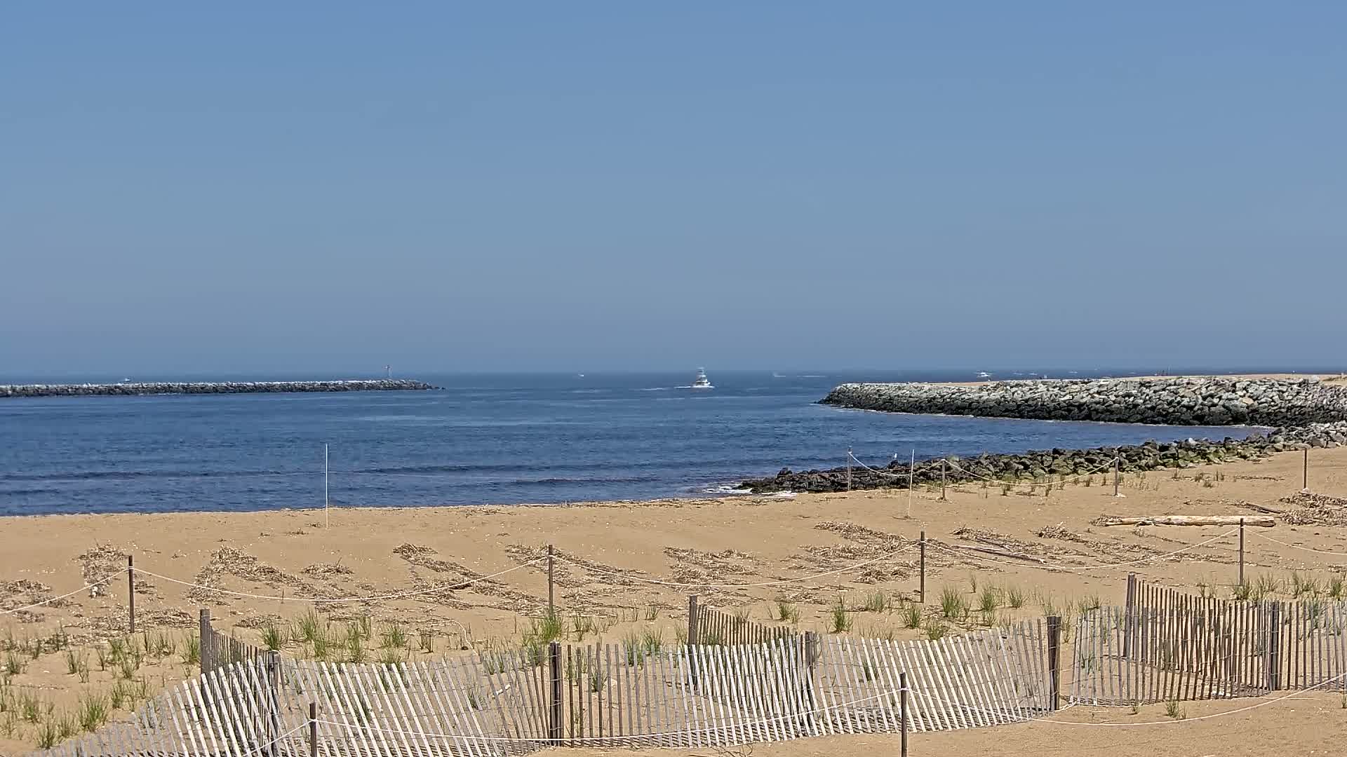A sandy beach with a wooden fence and small plants in the foreground overlooks a calm ocean inlet with rock jetties on either side, under a clear blue sky.