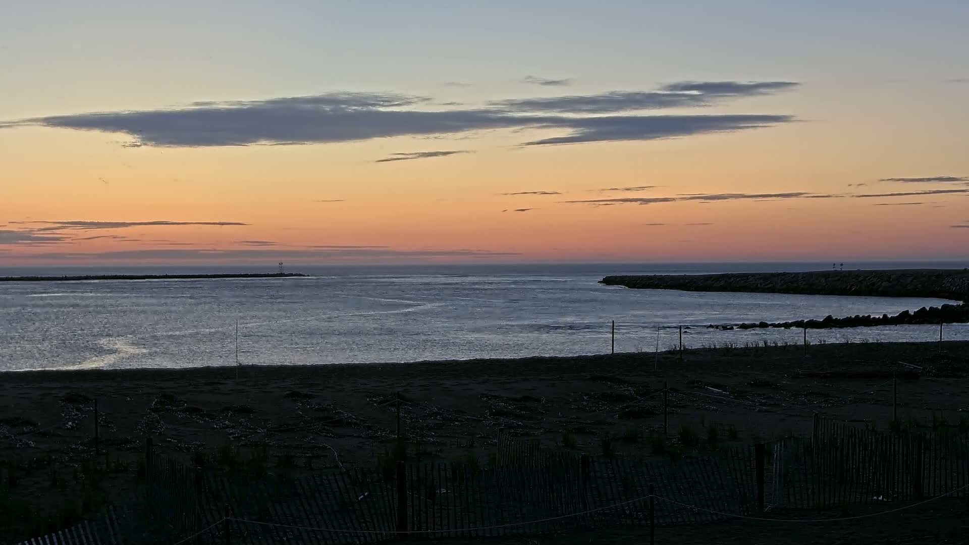 A calm ocean inlet at sunset, with a sandy beach and a dark fence in the foreground.