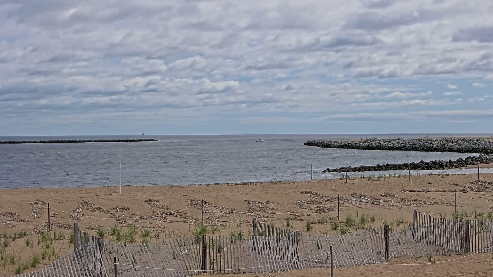 A sandy beach with a wooden fence and small plants, stretches to a calm body of water enclosed by rock jetties under a cloudy sky.