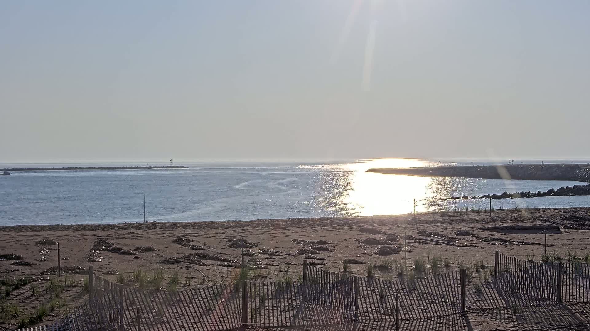 A sandy beach with a wooden fence is situated in front of a calm body of water that leads to a wider expanse of water under a bright, sunny sky.