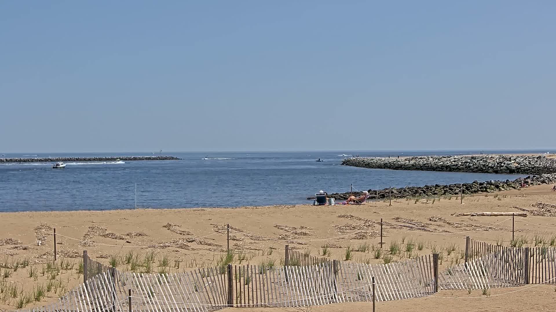 A sandy beach with a wooden fence and a few people relaxing is visible, overlooking a calm body of water with boats and rock jetties under a clear blue sky.