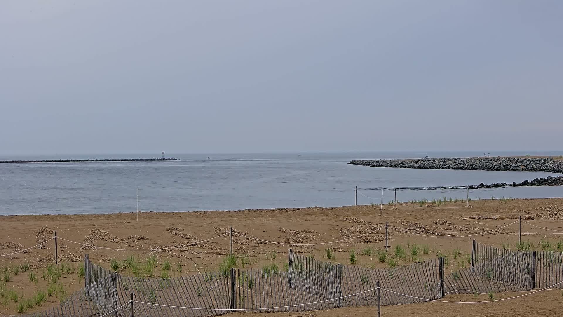 A sandy beach with small wooden fences protecting young vegetation stretches toward a calm body of water, bordered by rock jetties under an overcast sky.