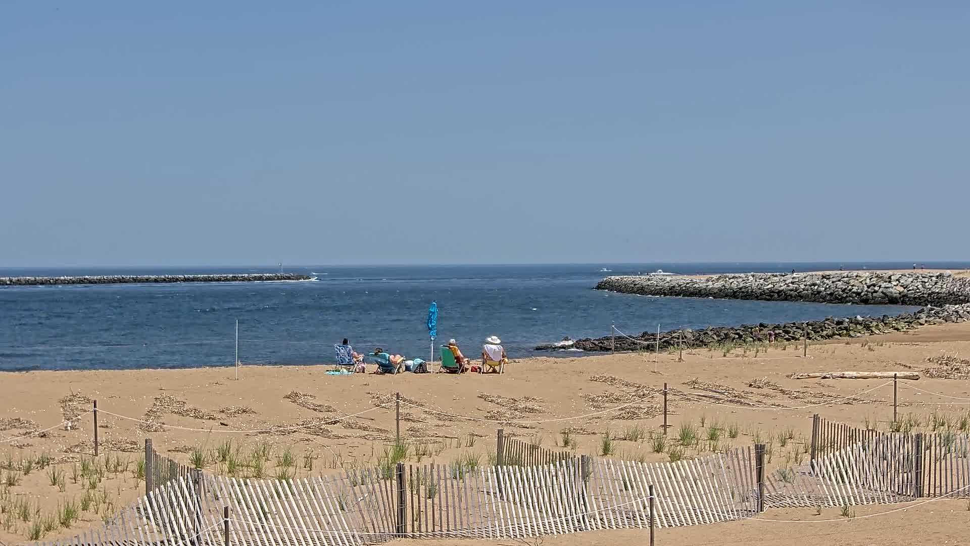 A sandy beach with a group of people sitting under an umbrella, separated from the ocean by a low wooden fence, on a clear sunny day.