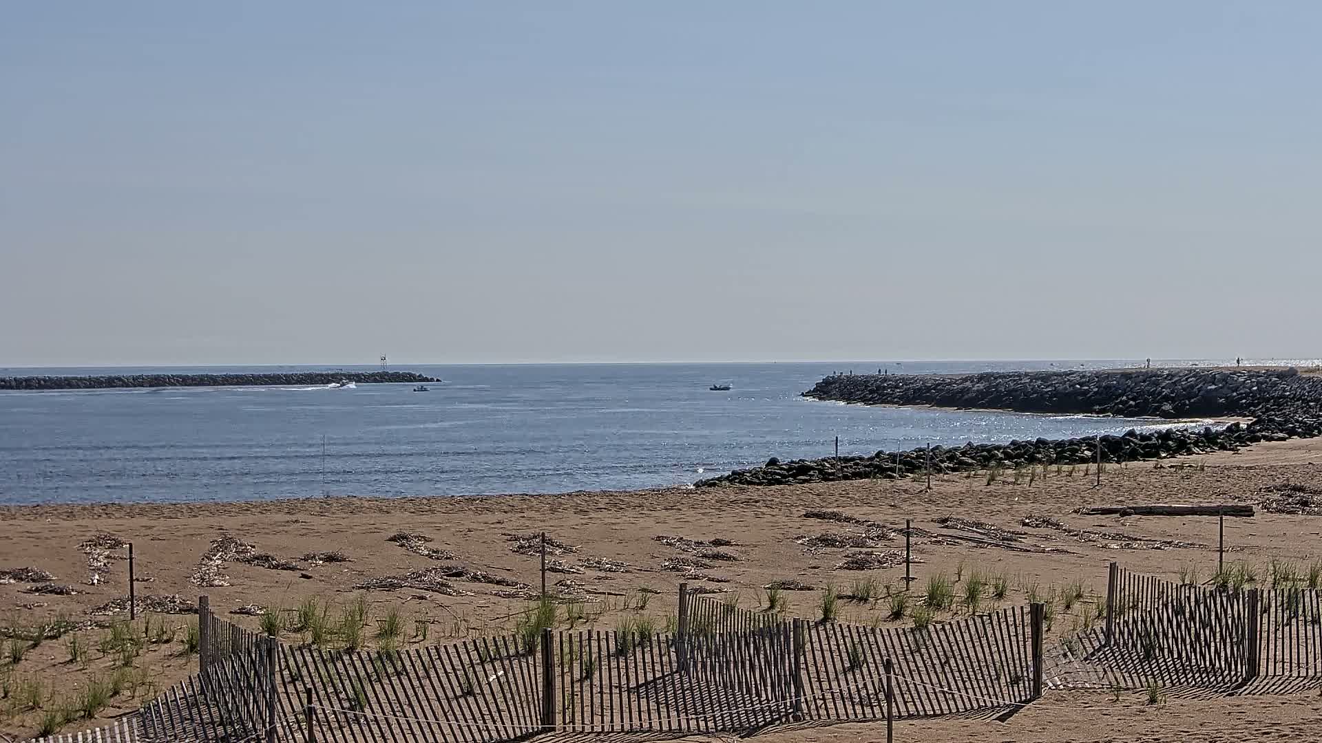 A sandy beach with a wooden fence stretches toward a calm ocean inlet, bookended by rock jetties, under a clear sky.