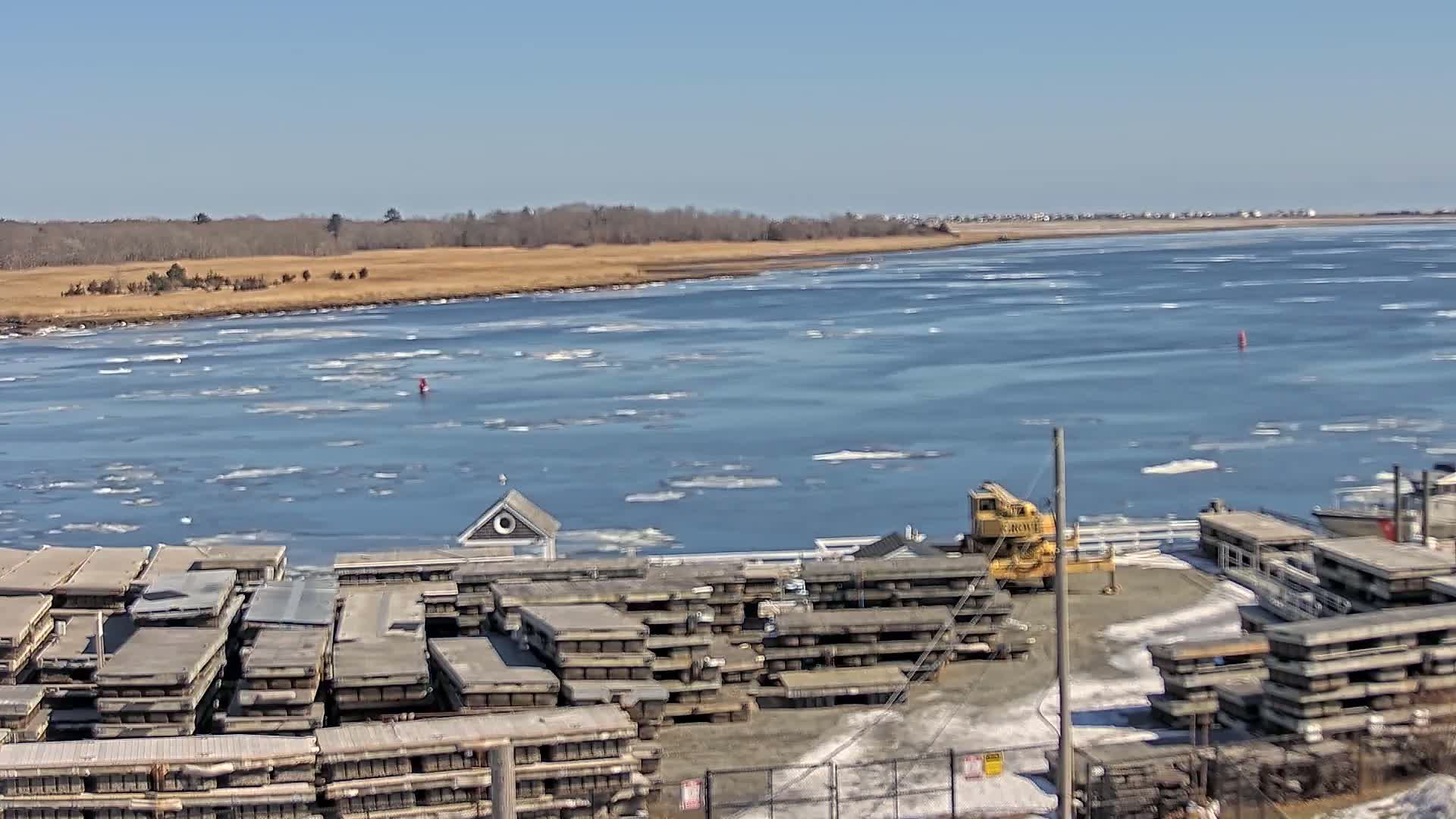 A partially ice-covered body of water is seen from a high vantage point, with a dock containing stacked planks and a crane in the foreground under a clear blue sky.